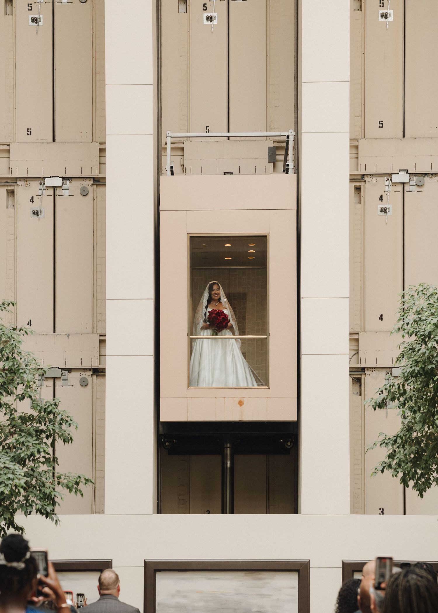 Bride in white gown and veil holding red bouquet stands in glass elevator of modern building during wedding.