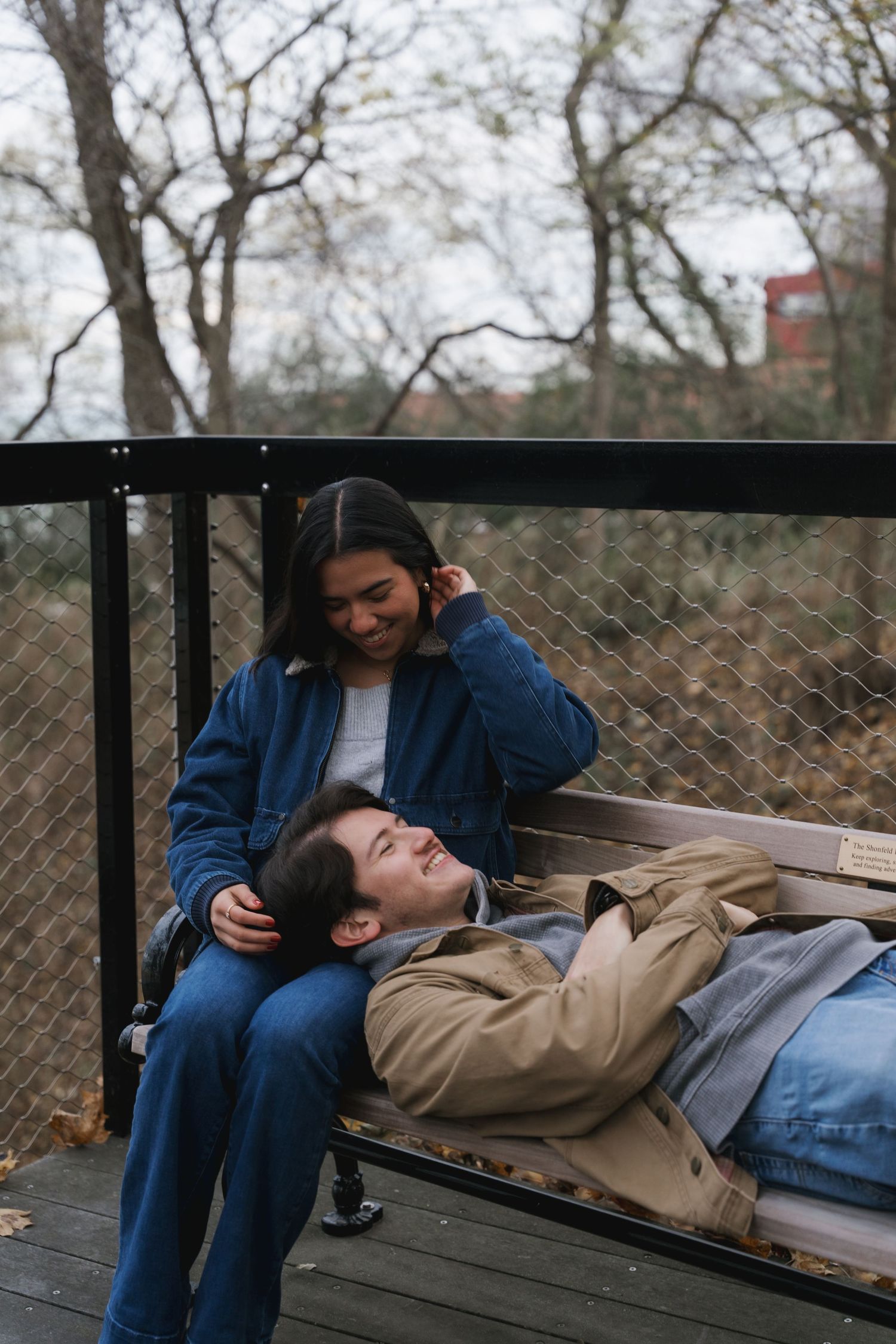 A couple relaxes on a park bench, woman smiling as man rests his head on her lap.