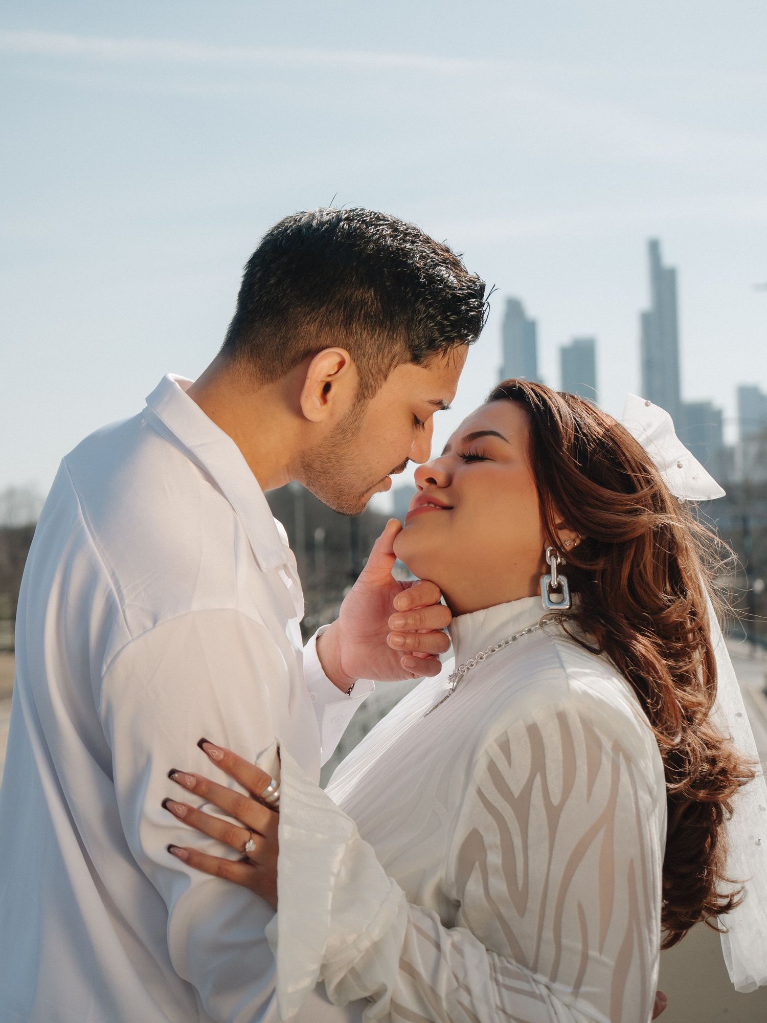 Couple in matching white outfits sharing a tender moment with a city skyline backdrop.