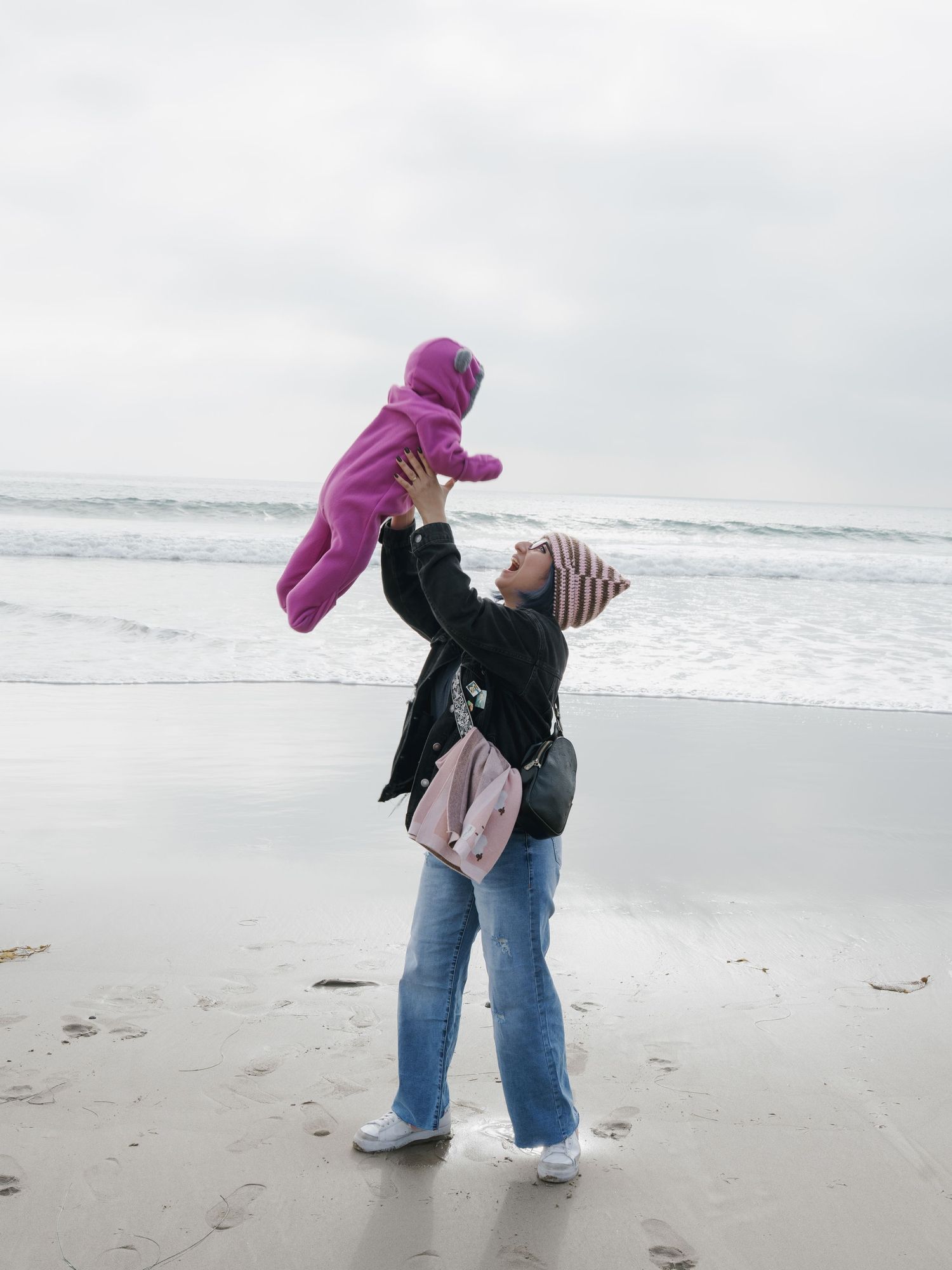 A woman lifts a baby in a pink snowsuit high above her head on a cloudy beach near ocean waves.