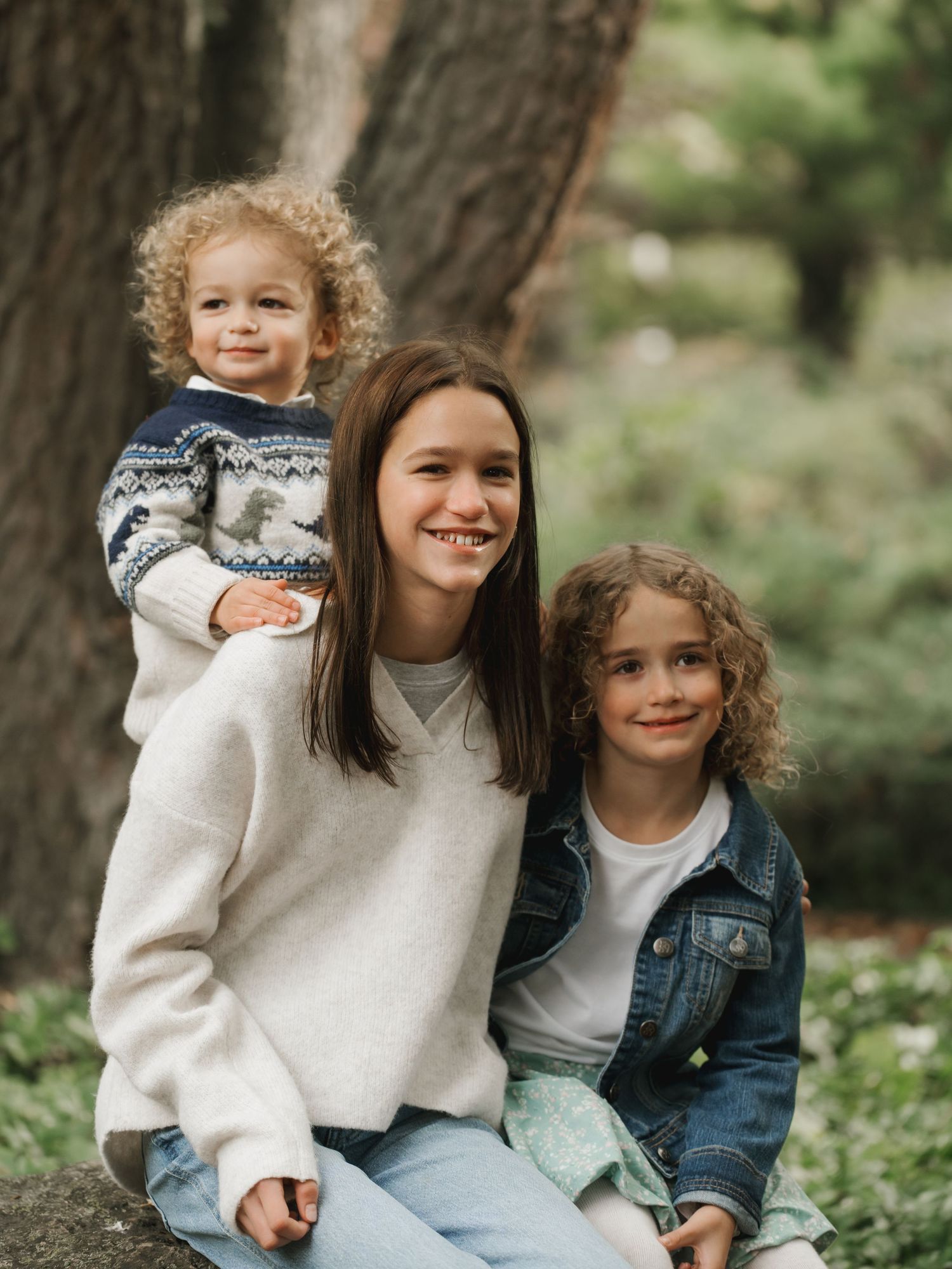 A young woman smiles with two curly-haired children in a forest setting, wearing cozy sweaters.