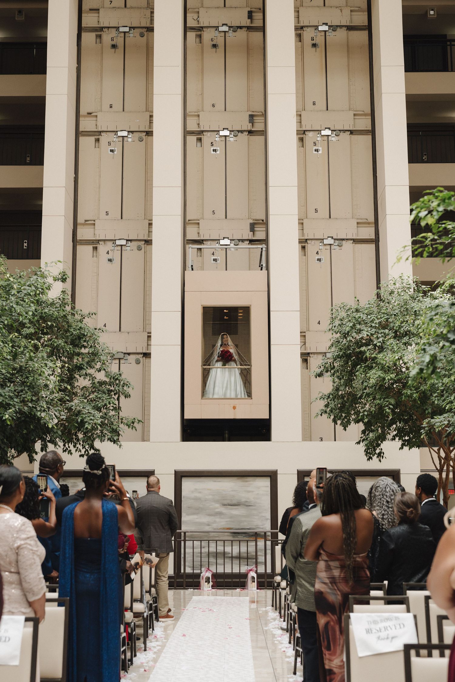 Bride in white gown descends glass elevator at indoor atrium wedding ceremony with seated guests.