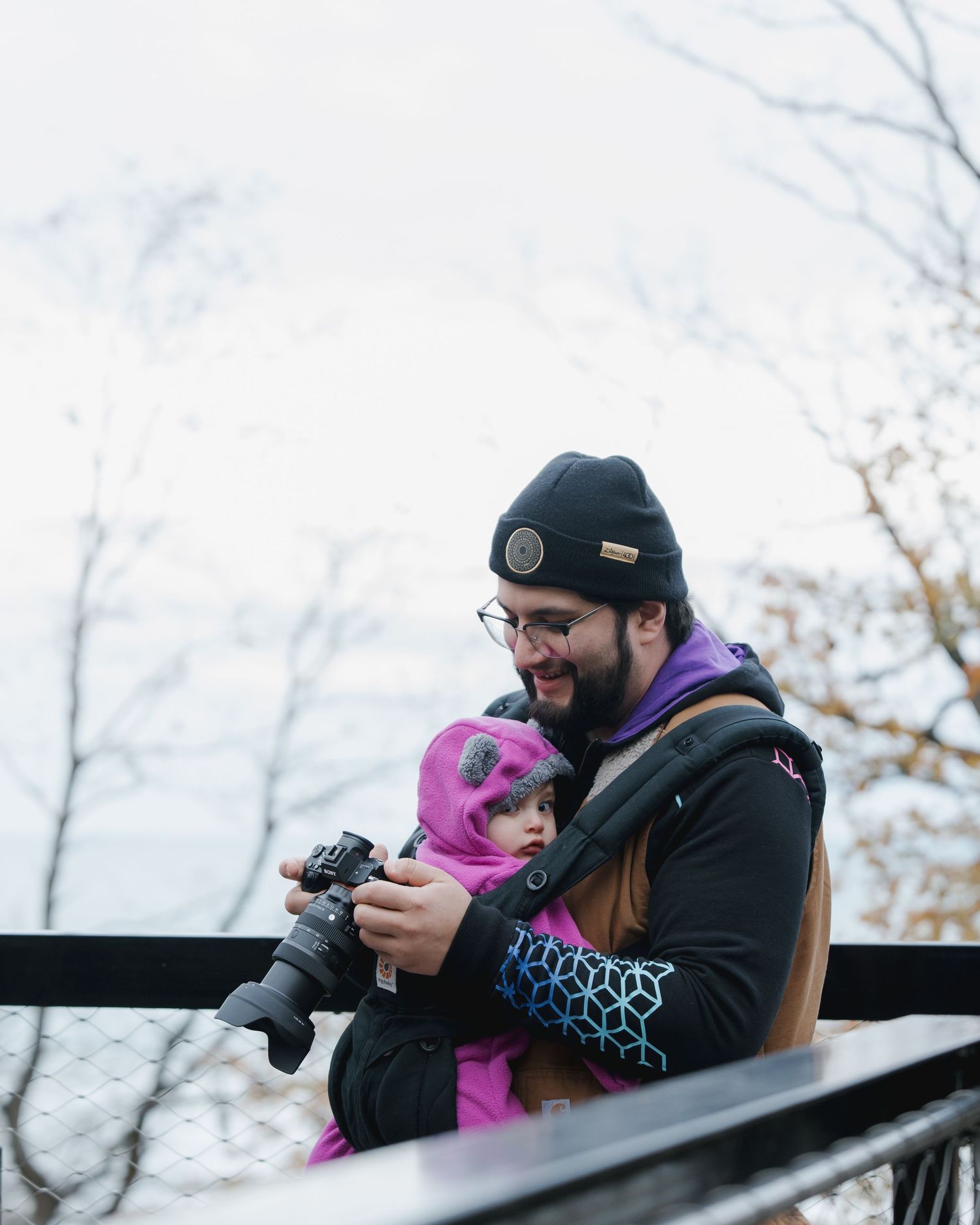A bearded man in a black beanie holds a camera and a baby in a pink snowsuit on an outdoor overlook.