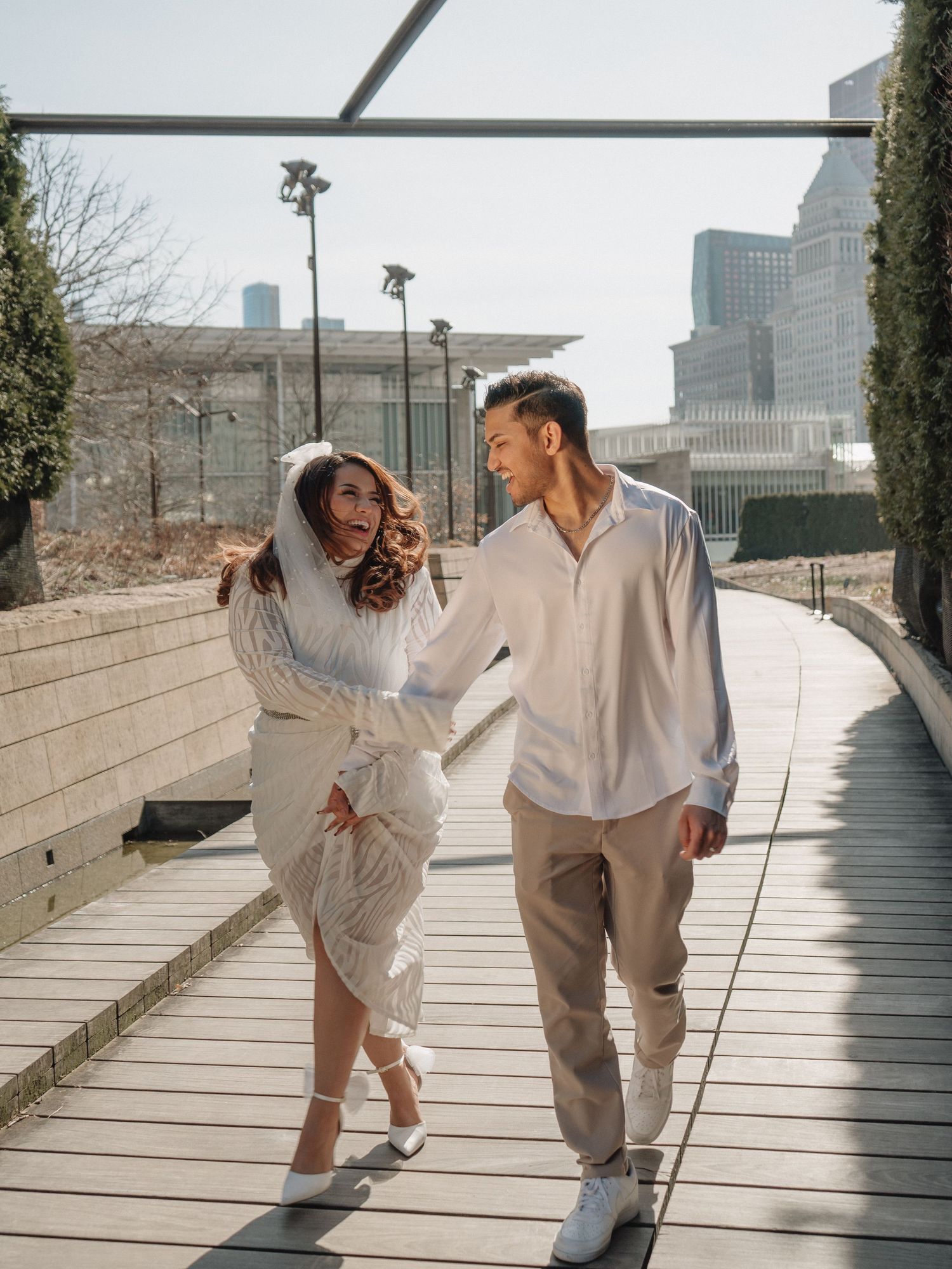 Joyful couple in white outfits walking and laughing together on a sunny urban boardwalk.
