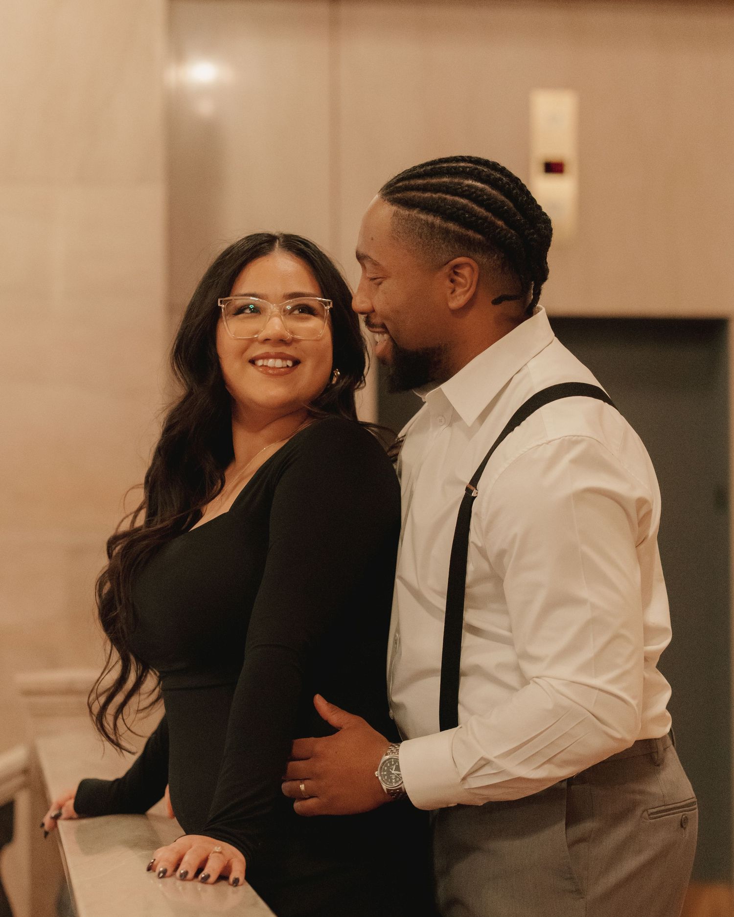Smiling couple posing together, woman in black outfit with glasses, man in white shirt with suspenders.