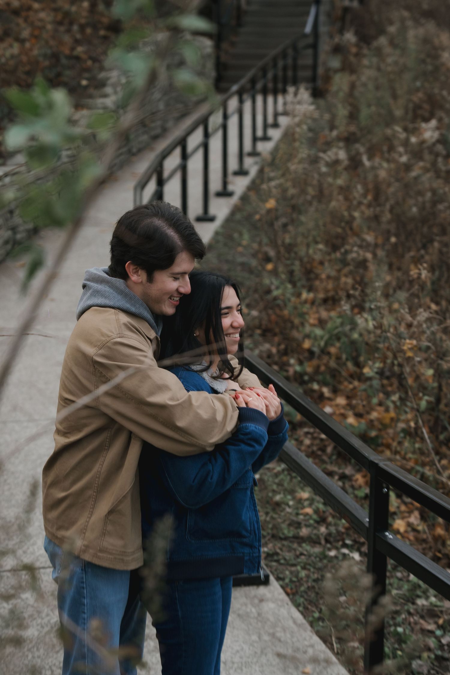 A young couple embraces and smiles while standing on outdoor stairs with autumn foliage.