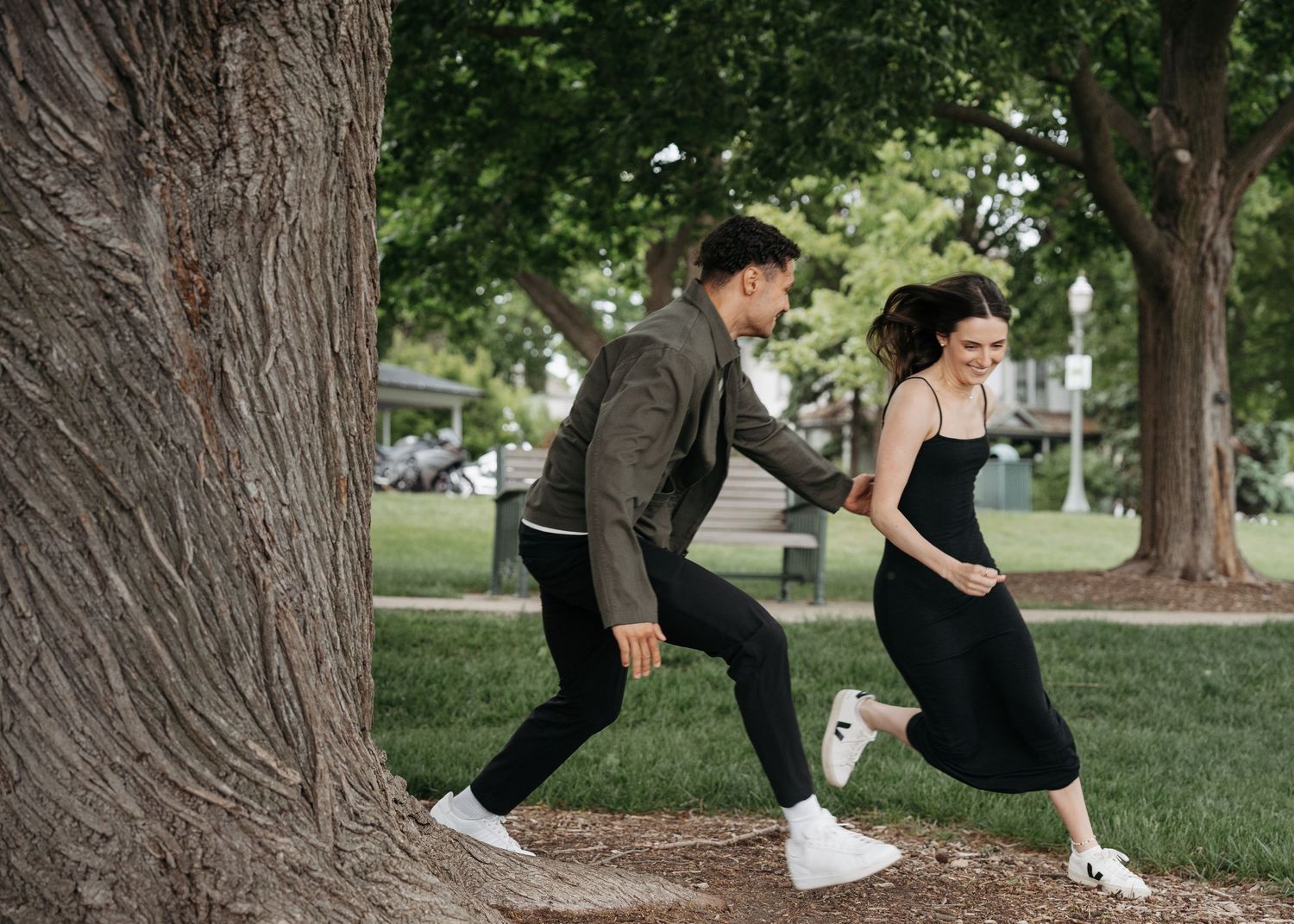 A young couple playfully runs around a large tree in a lush green park on a sunny day.