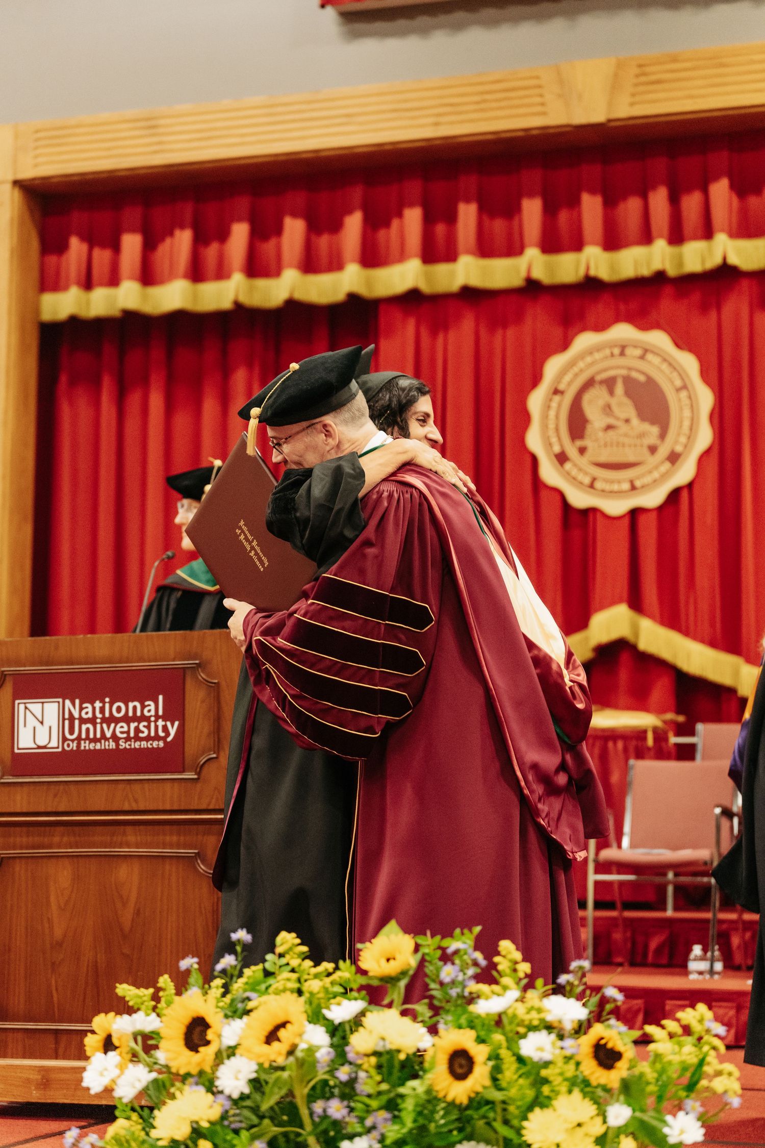 Two graduates in academic regalia share a hug at National University of Health Sciences commencement ceremony.