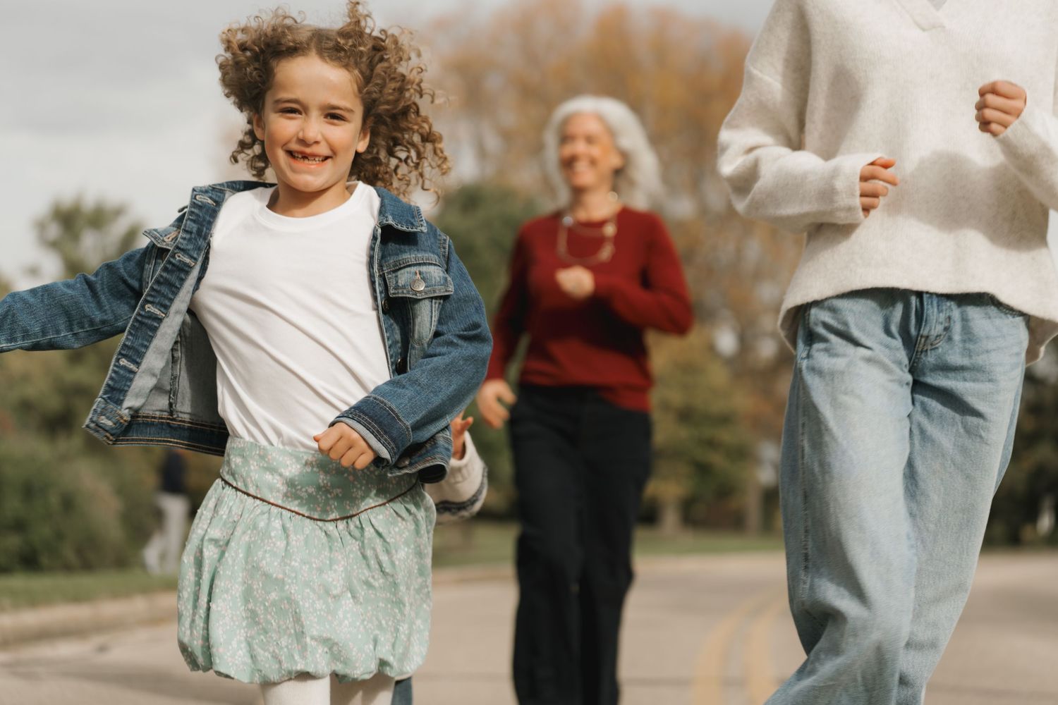 Young girl in denim jacket running outdoors with family in background.