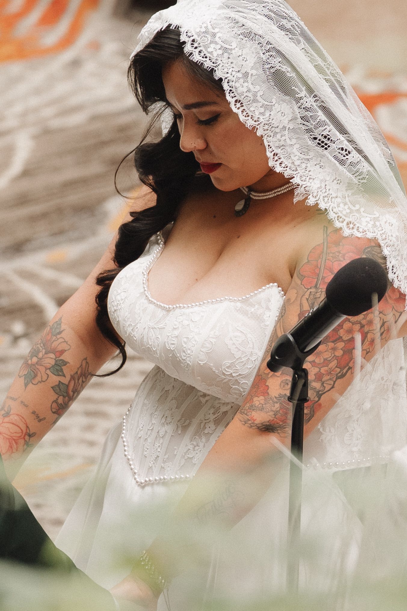 Tattooed bride in white lace gown and veil stands at microphone during outdoor wedding ceremony.