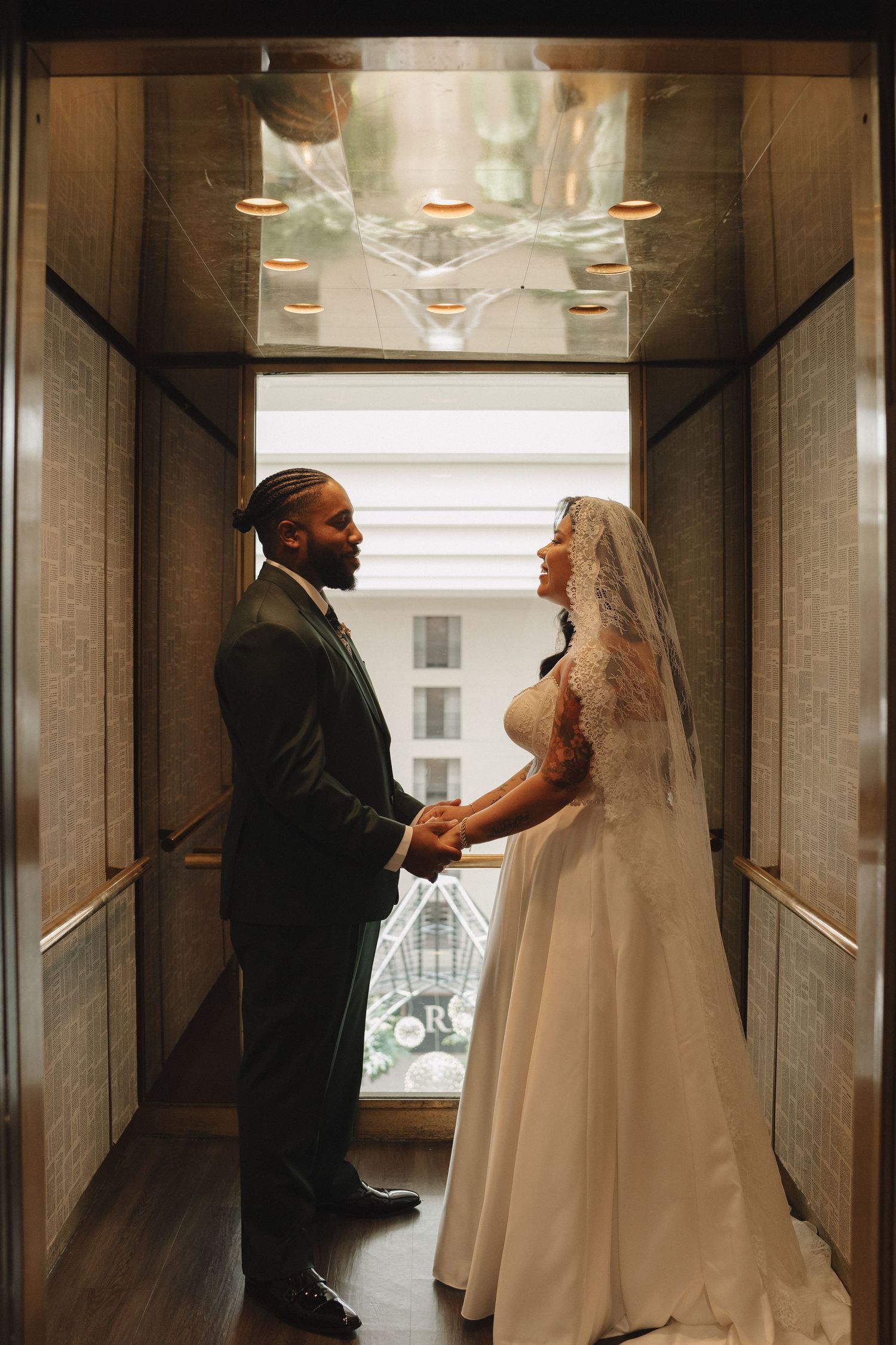 Bride and groom hold hands facing each other in an elegant elevator, she in a lace gown with veil, he in a dark suit.