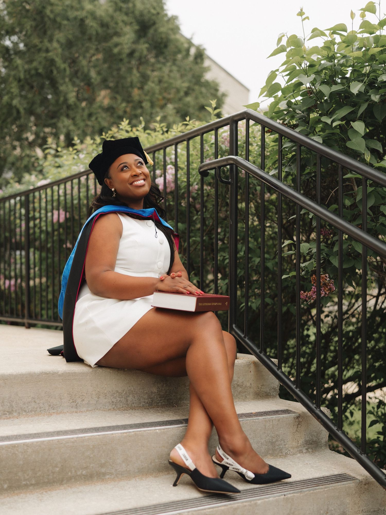 Smiling graduate in cap and gown sitting on stone steps holding a book, wearing a white dress and heels.