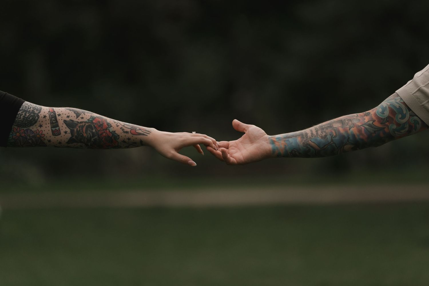 Two tattooed arms reaching toward each other with fingertips almost touching against a blurred green background.