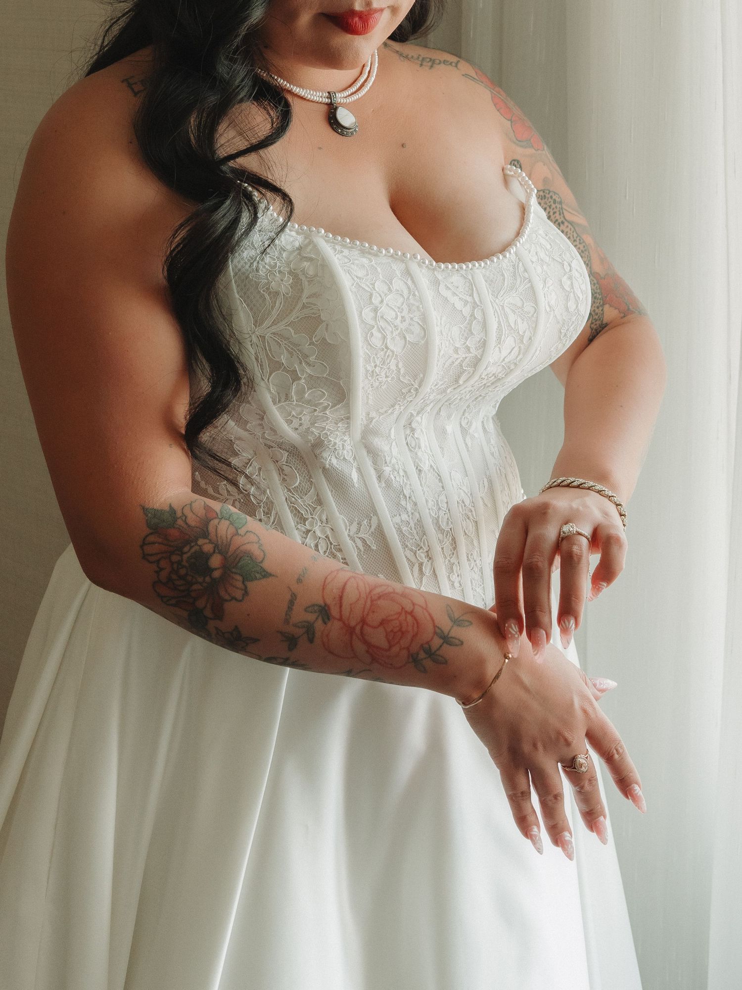 Bride in white lace corset wedding gown with floral tattoos on arms, wearing pearl necklace and bracelet.