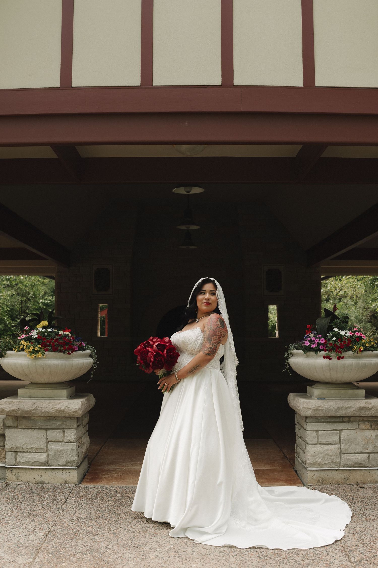Bride in white lace gown and veil holds red bouquet, posing before a building entrance with floral arrangements.