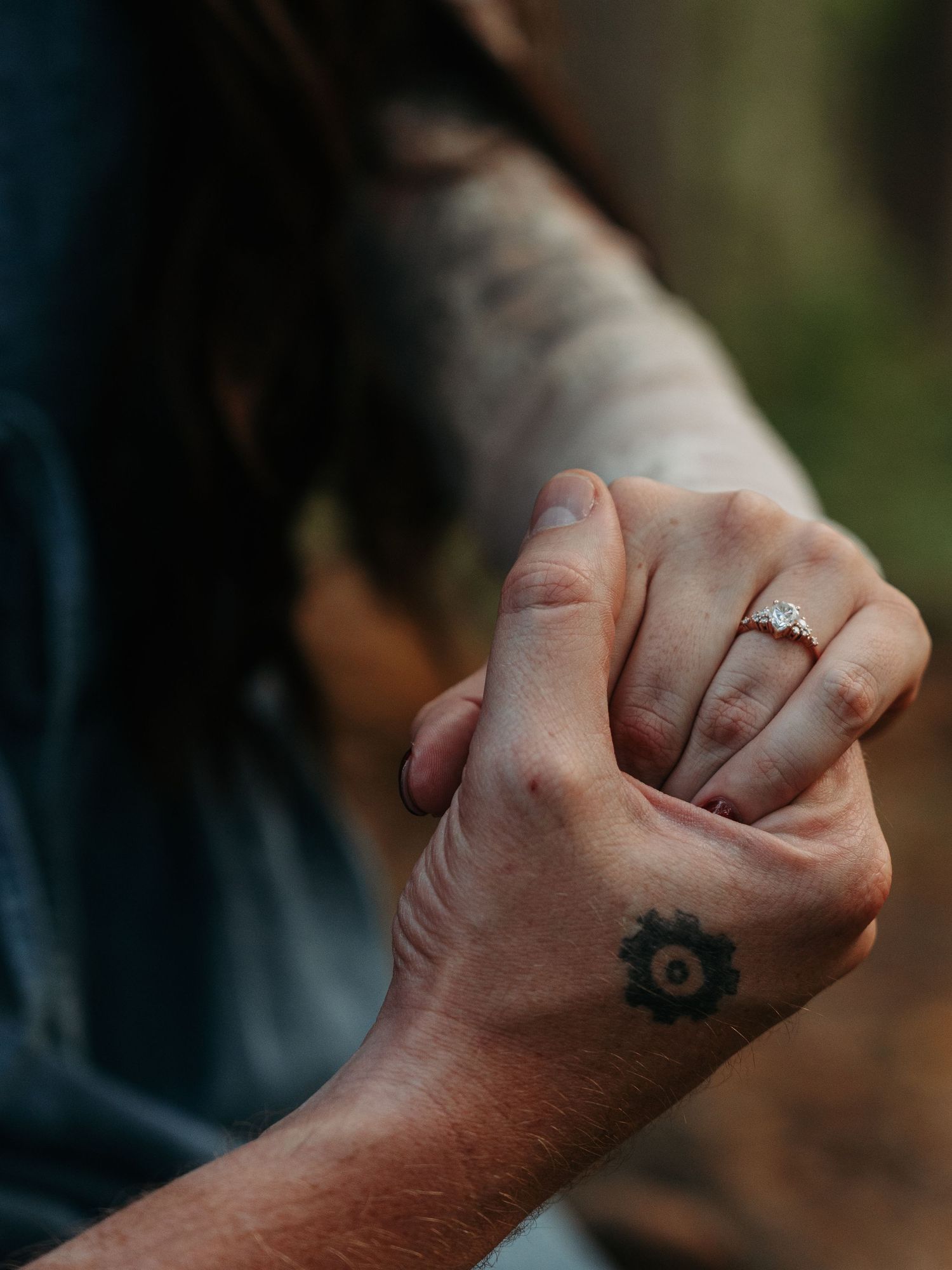 Close-up of two hands intertwined, one wearing an engagement ring and the other with a gear tattoo.
