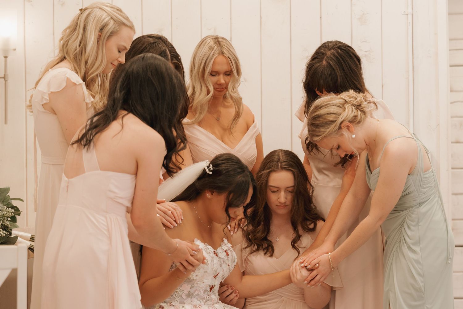 Bridesmaids in blush dresses gather around bride in circle for prayer before wedding ceremony.