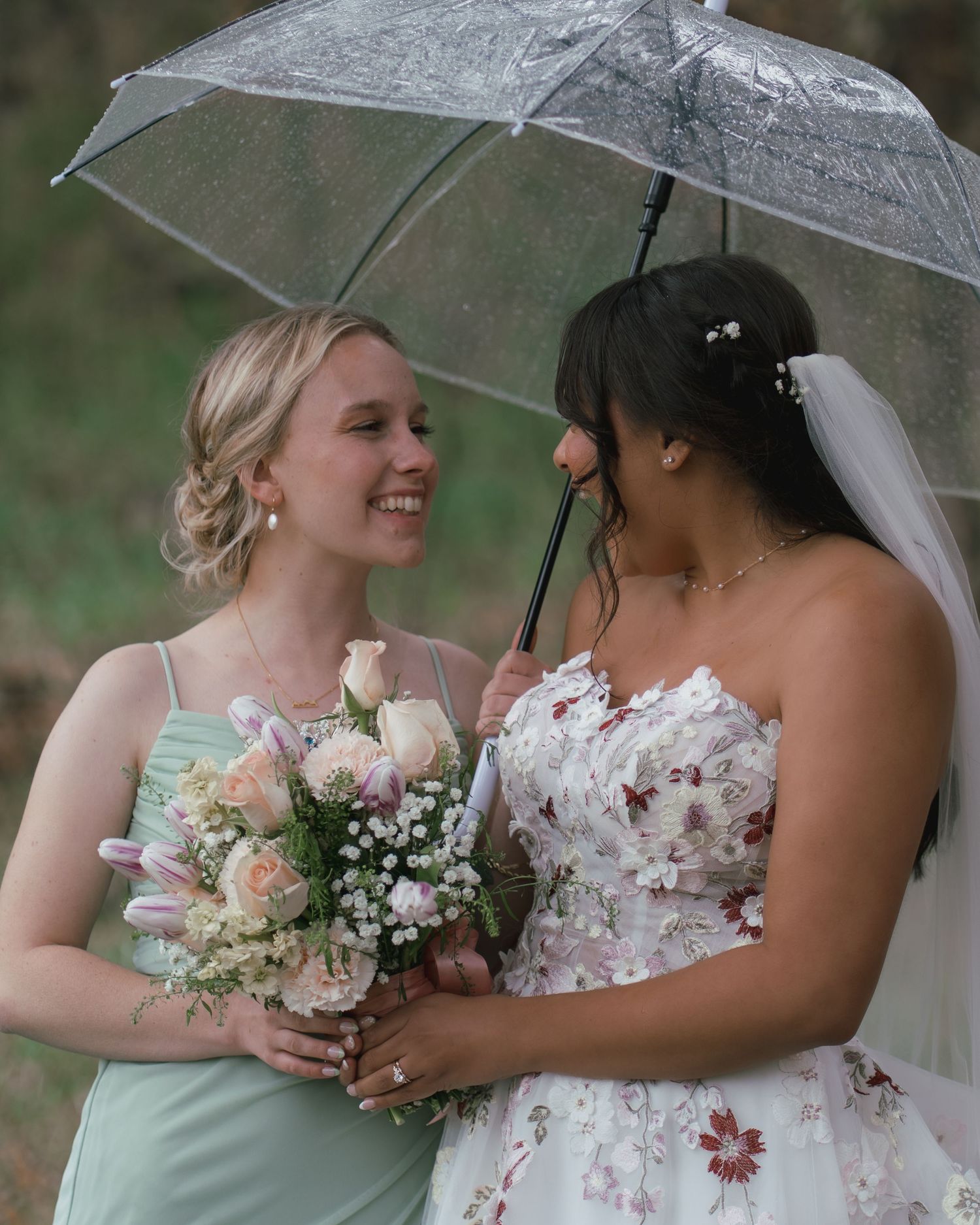 Bride in floral gown and bridesmaid in sage green dress smile under umbrella in the rain.