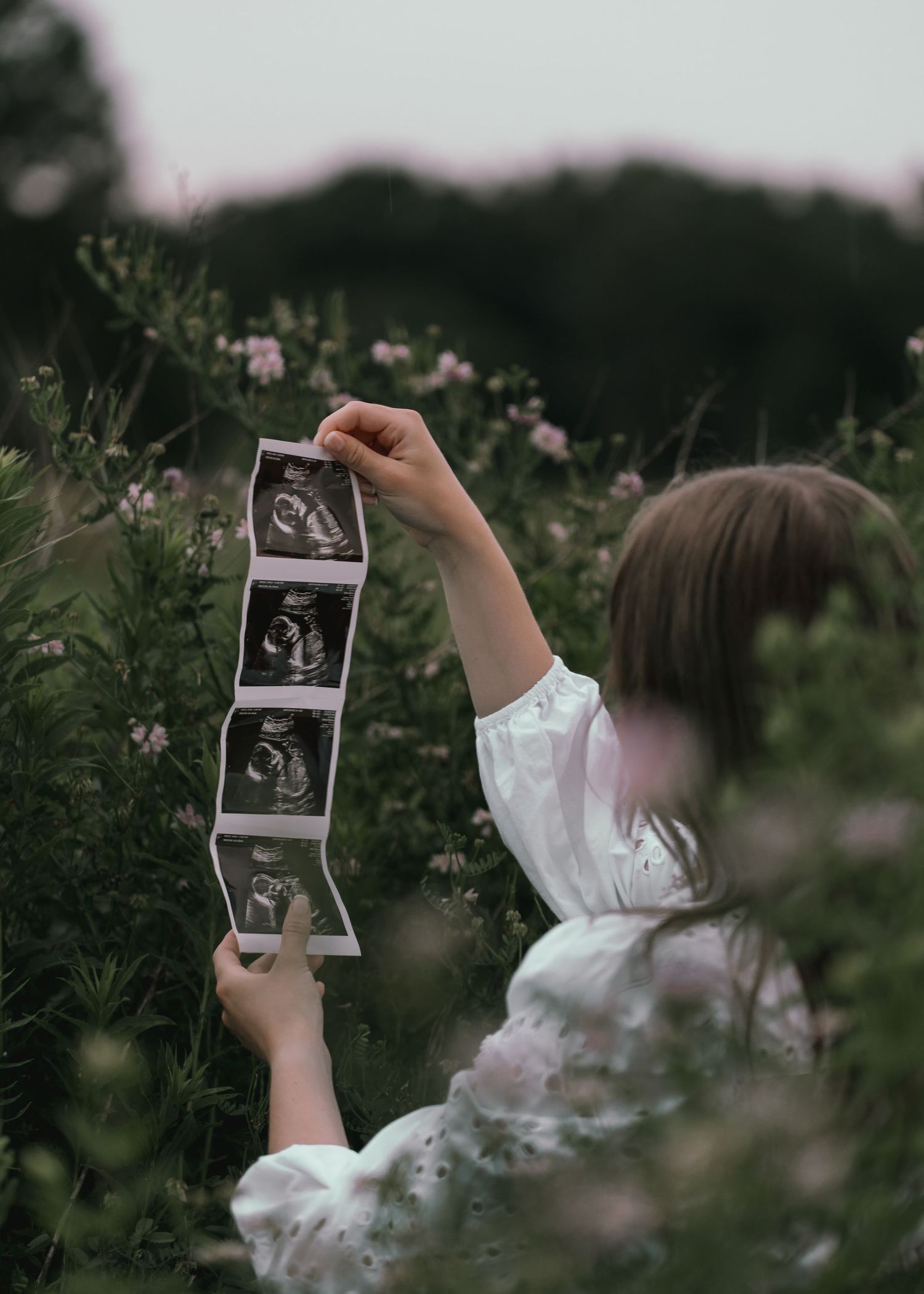 Mother holds ultrasound photos up among lush garden greenery and pink flowers.