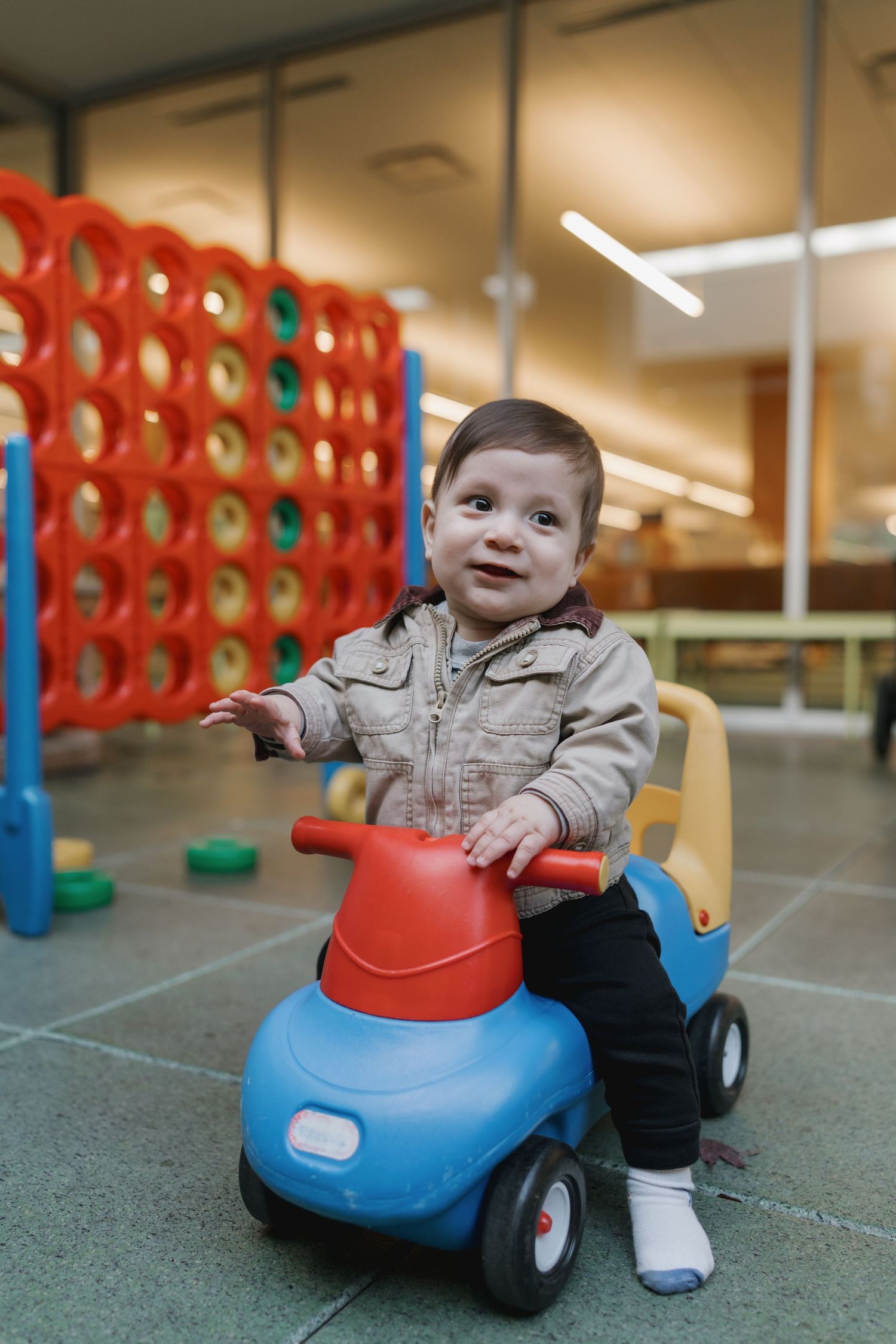 Smiling baby boy sitting on a blue and red toy ride-on car in a colorful indoor play area.