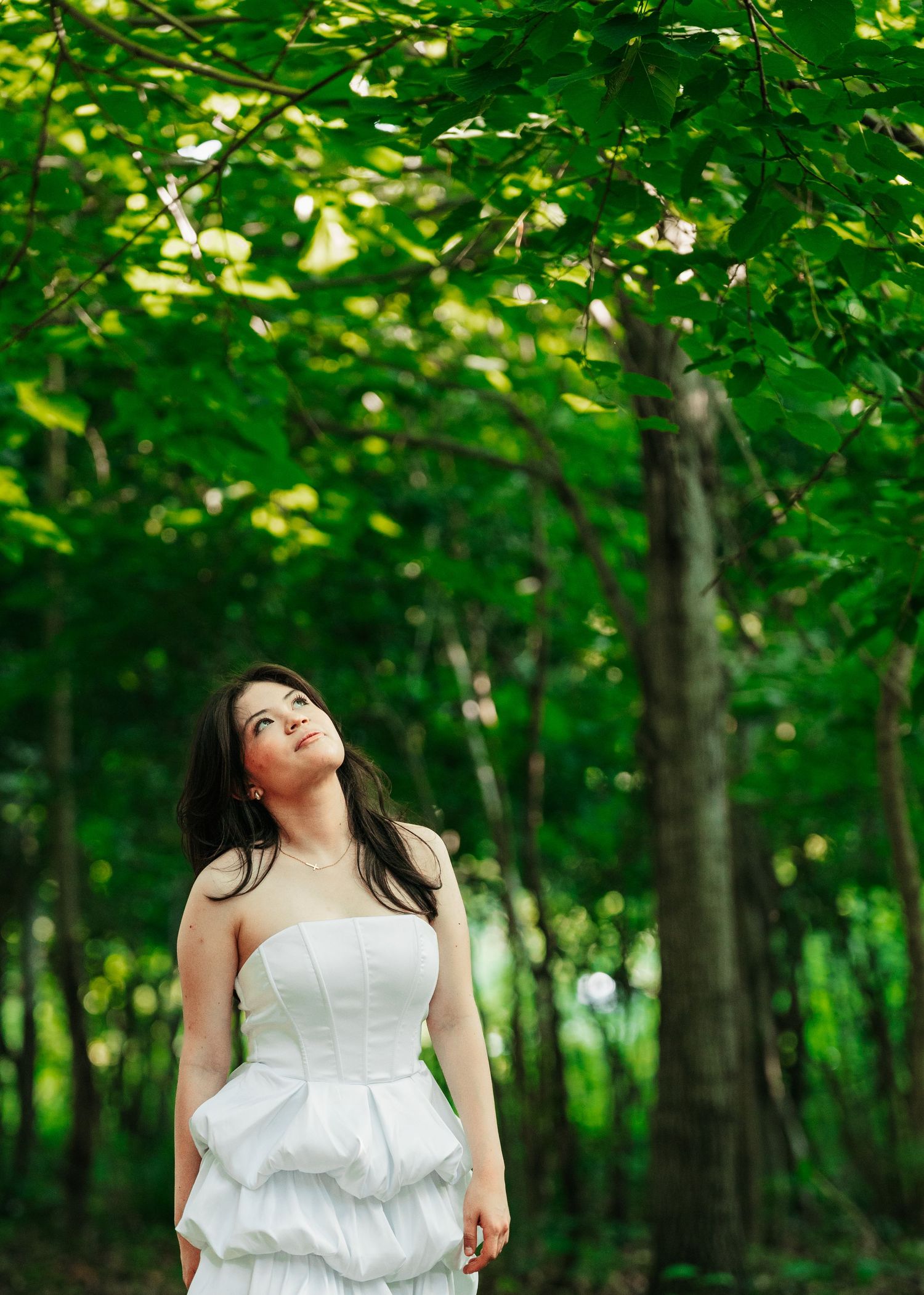 Young woman in white strapless tiered dress looking up among lush green trees outdoors.