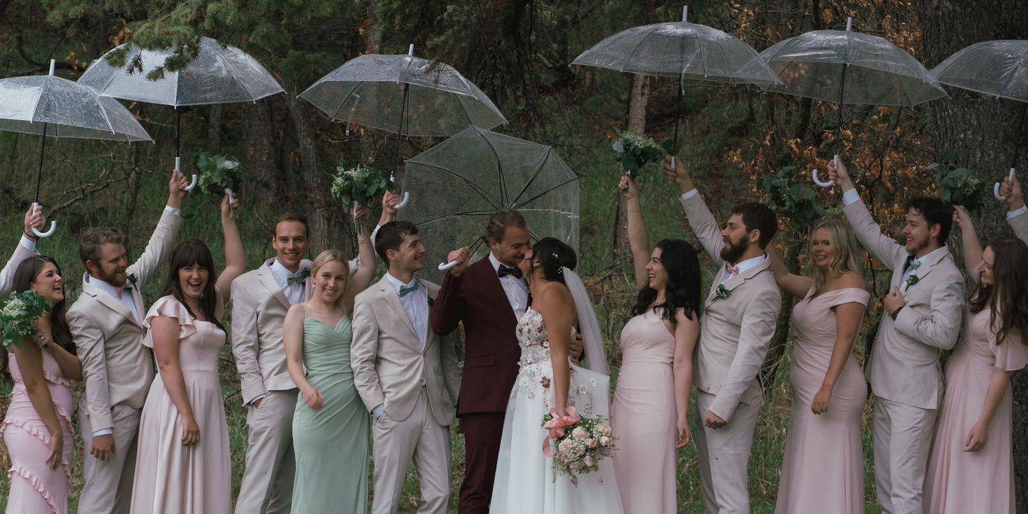 Wedding party raises clear umbrellas in the rain during an outdoor ceremony in a wooded setting.