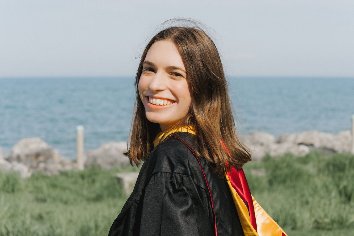 Smiling young woman in black and red graduation gown posing outdoors near rocky shoreline on clear day.