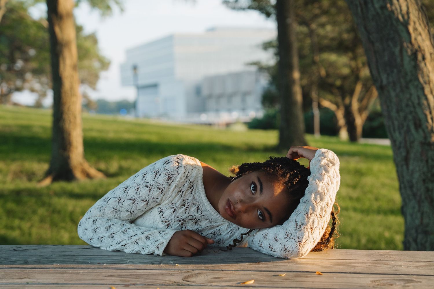 Young Black girl resting her head on a wooden table outdoors, wearing a white knit sweater in a park setting.