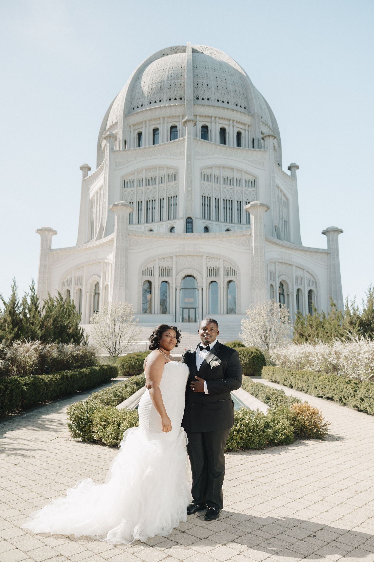 A bride in a ruffled mermaid-style gown and a groom in a classic tuxedo pose on a sunlit path during their outdoor wedding celebration.