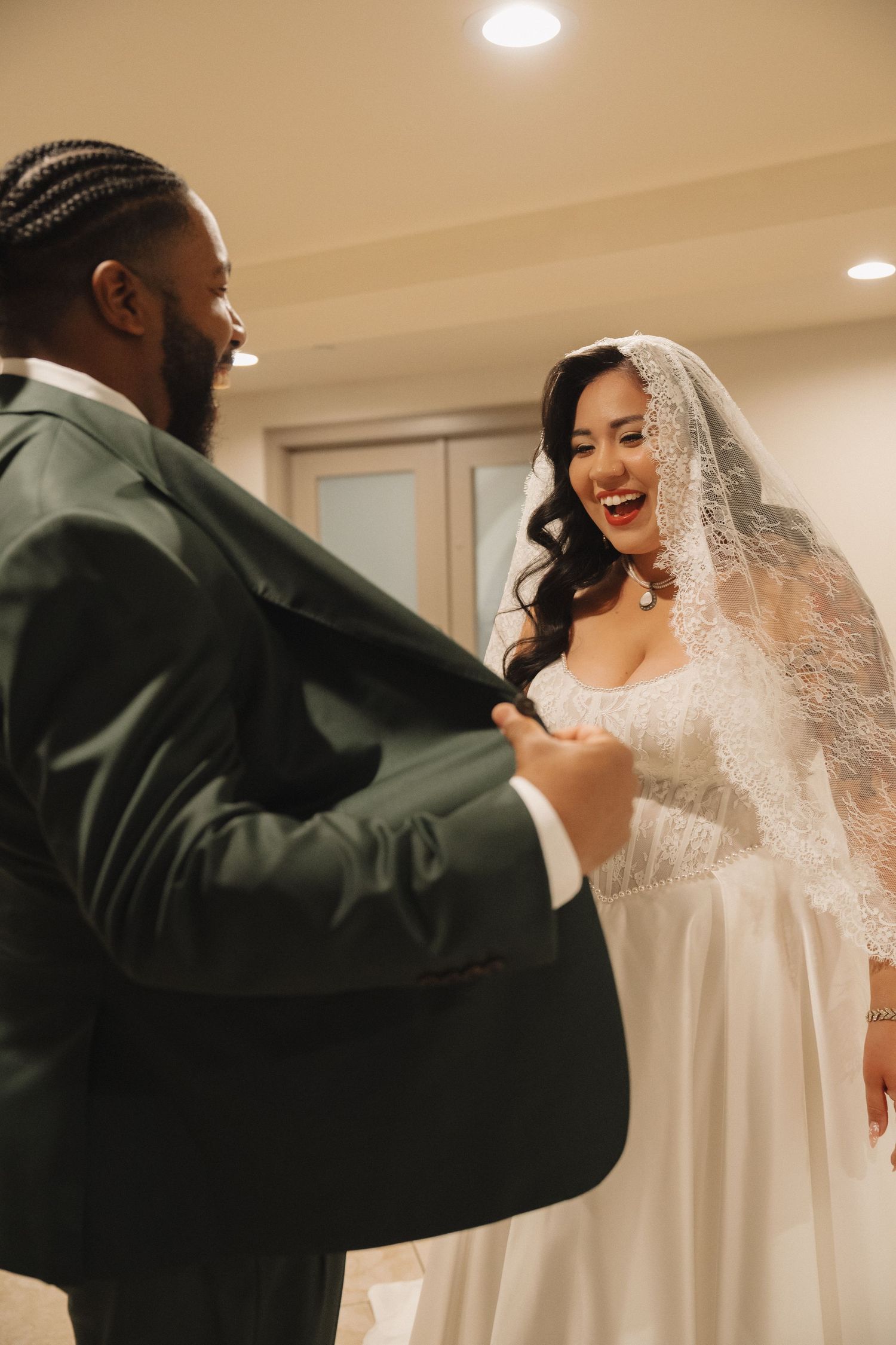 A joyful bride in a lace veil laughs with her groom in a green suit during their wedding day first look.
