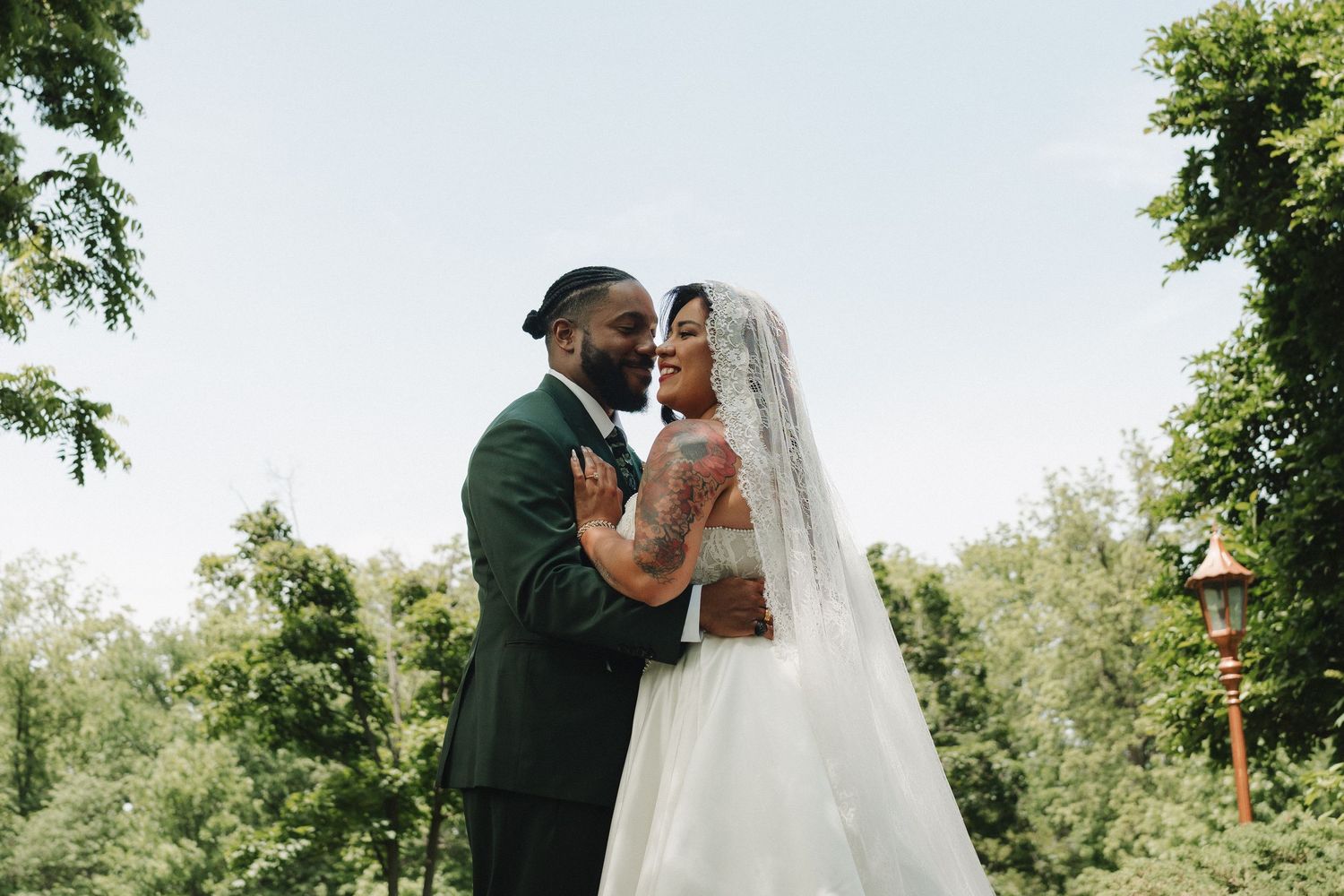 A joyful newlywed couple embraces outdoors, bride in white gown with veil, groom in dark suit, surrounded by lush greenery.