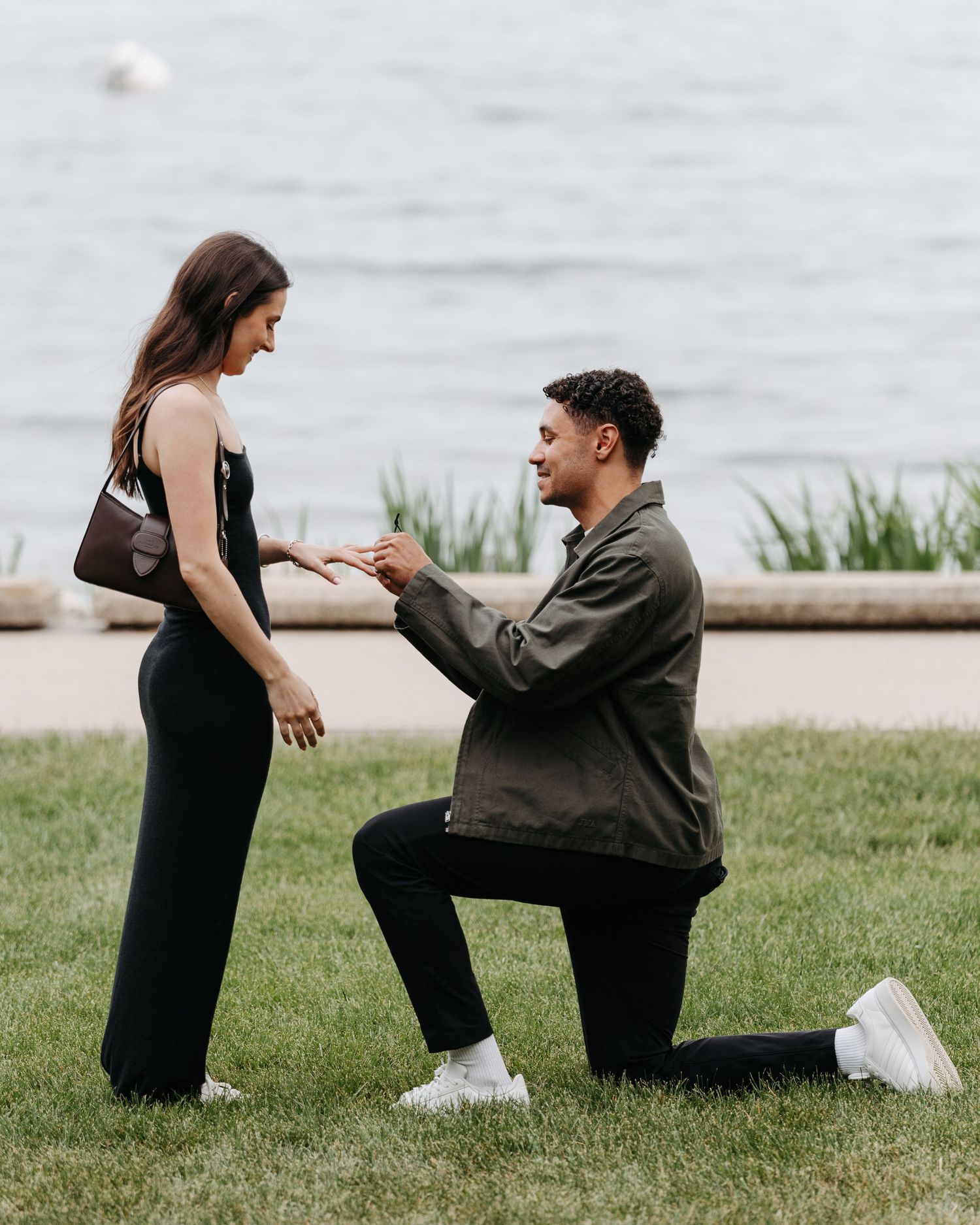 Man proposes on one knee to woman in black dress near waterfront on green grass, holding her hand.