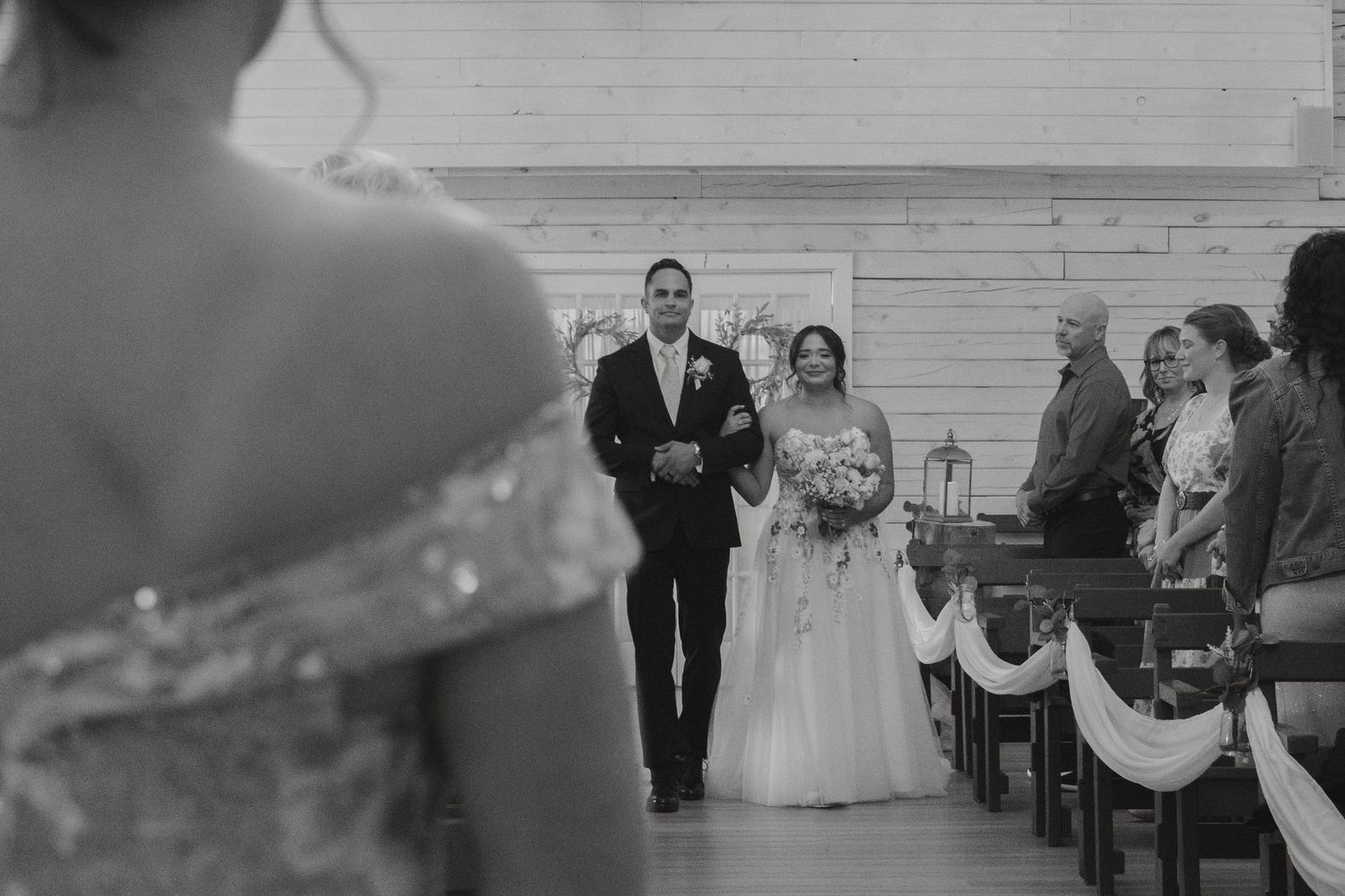 Black and white wedding photo of bride walking down the aisle with escort toward waiting groom in rustic chapel.