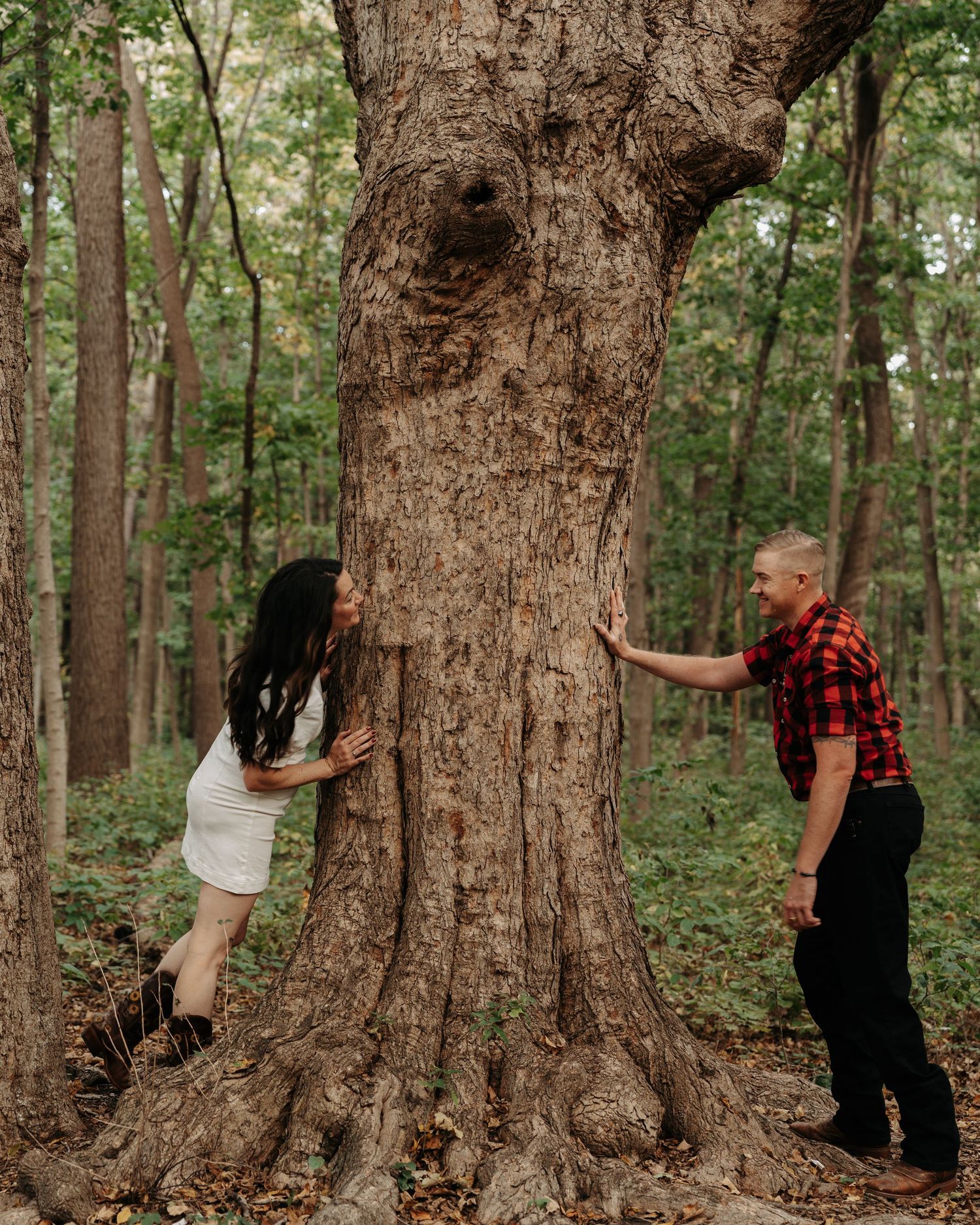 Playful couple peeking around opposite sides of a massive tree trunk in a lush forest.