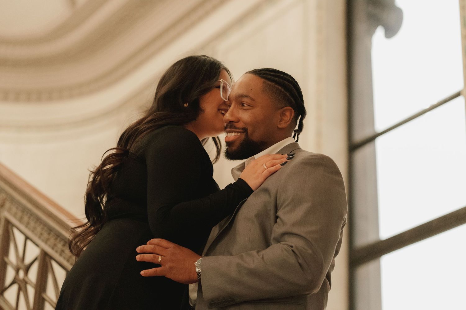 Joyful couple embracing and smiling near elegant staircase in bright natural light.