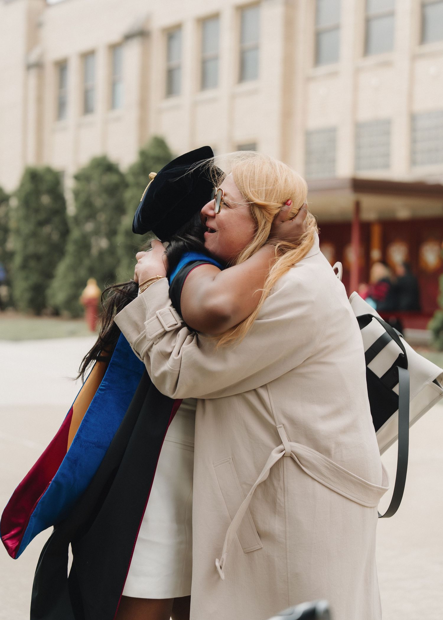 Graduate in cap and gown shares a warm hug with a proud family member at an outdoor university commencement ceremony.