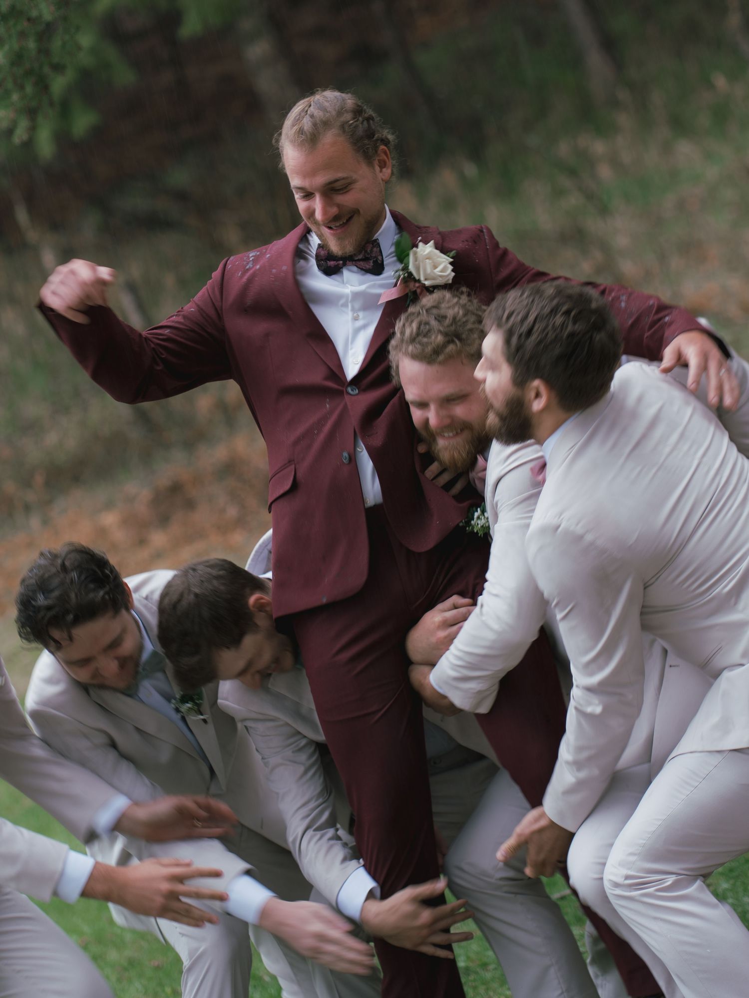 Groomsmen playfully lift the groom in a burgundy suit during fun outdoor wedding party photos.