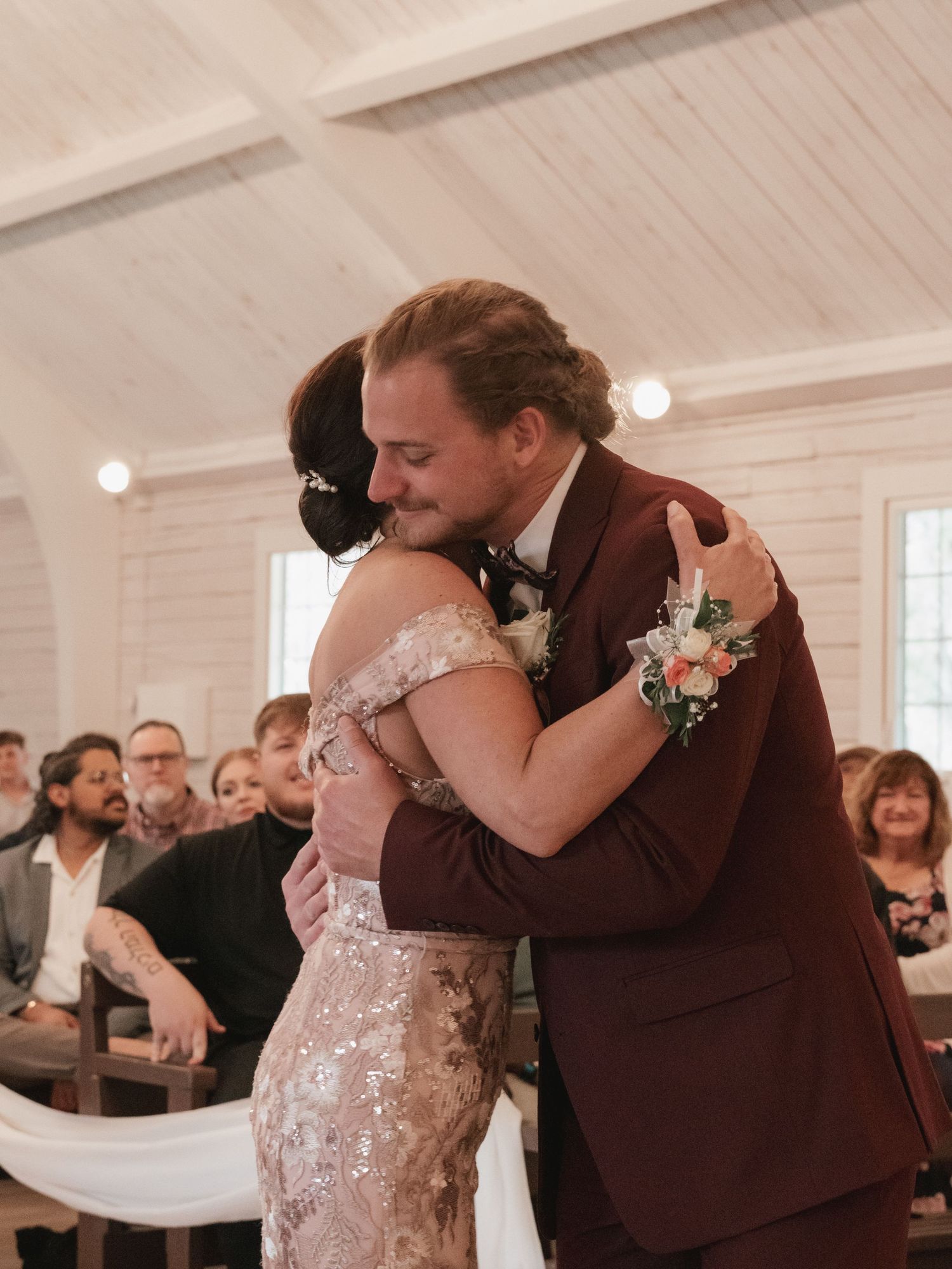 Groom in burgundy suit shares emotional hug with mother of the bride in sequin dress at wedding ceremony.