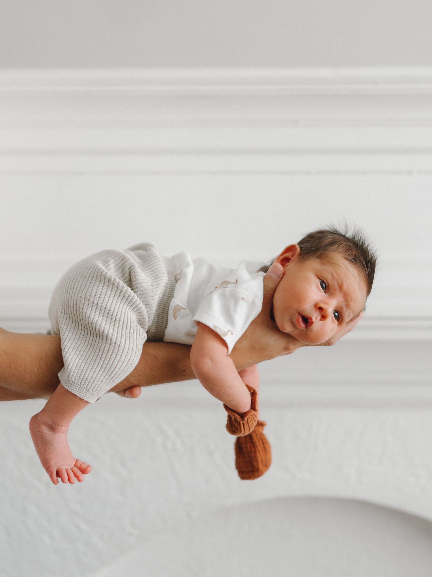 Newborn baby in white onesie and beige pants held safely in parent's hands against white wall.