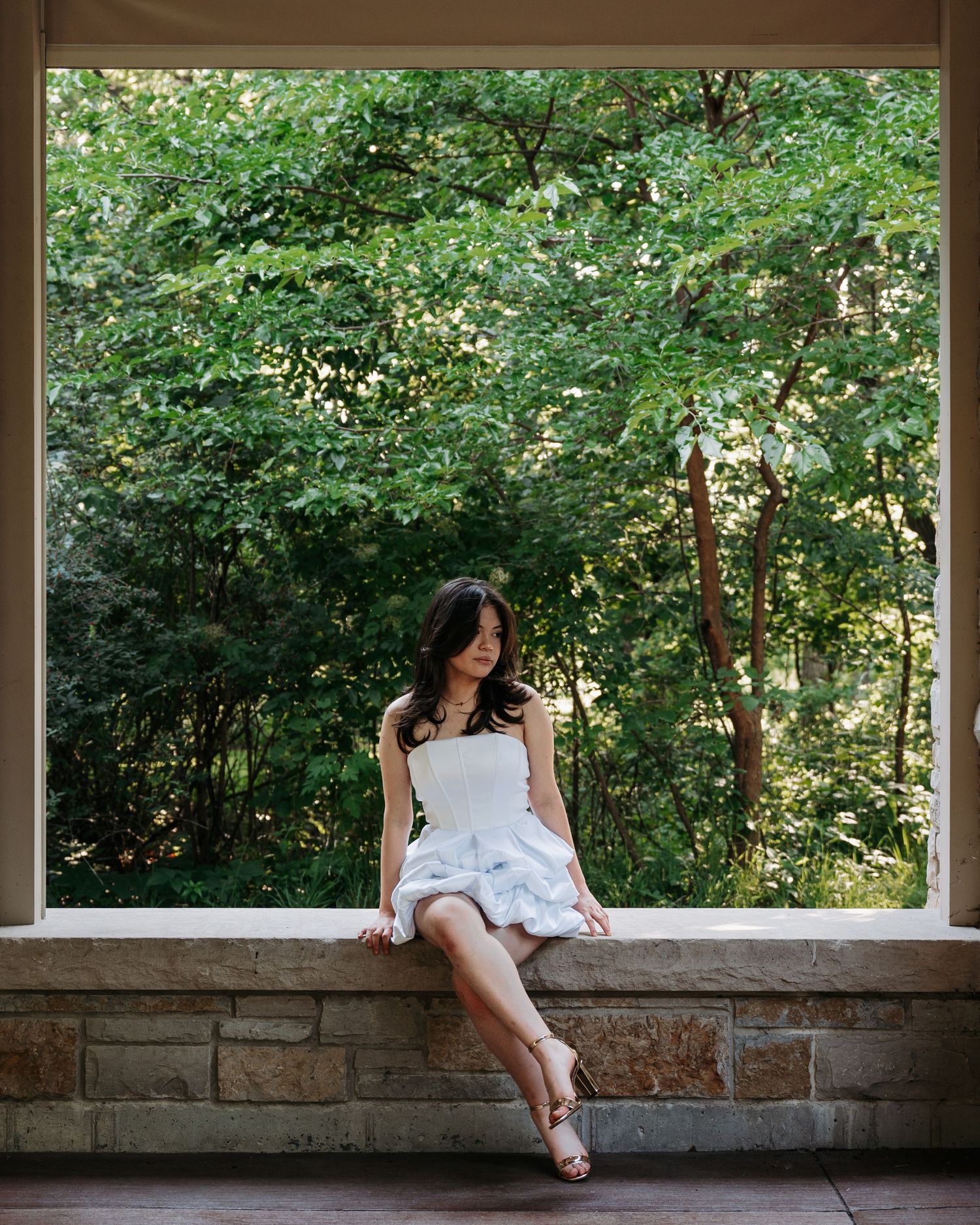 Young woman in white ruffled dress sitting on stone ledge before large window with green trees.