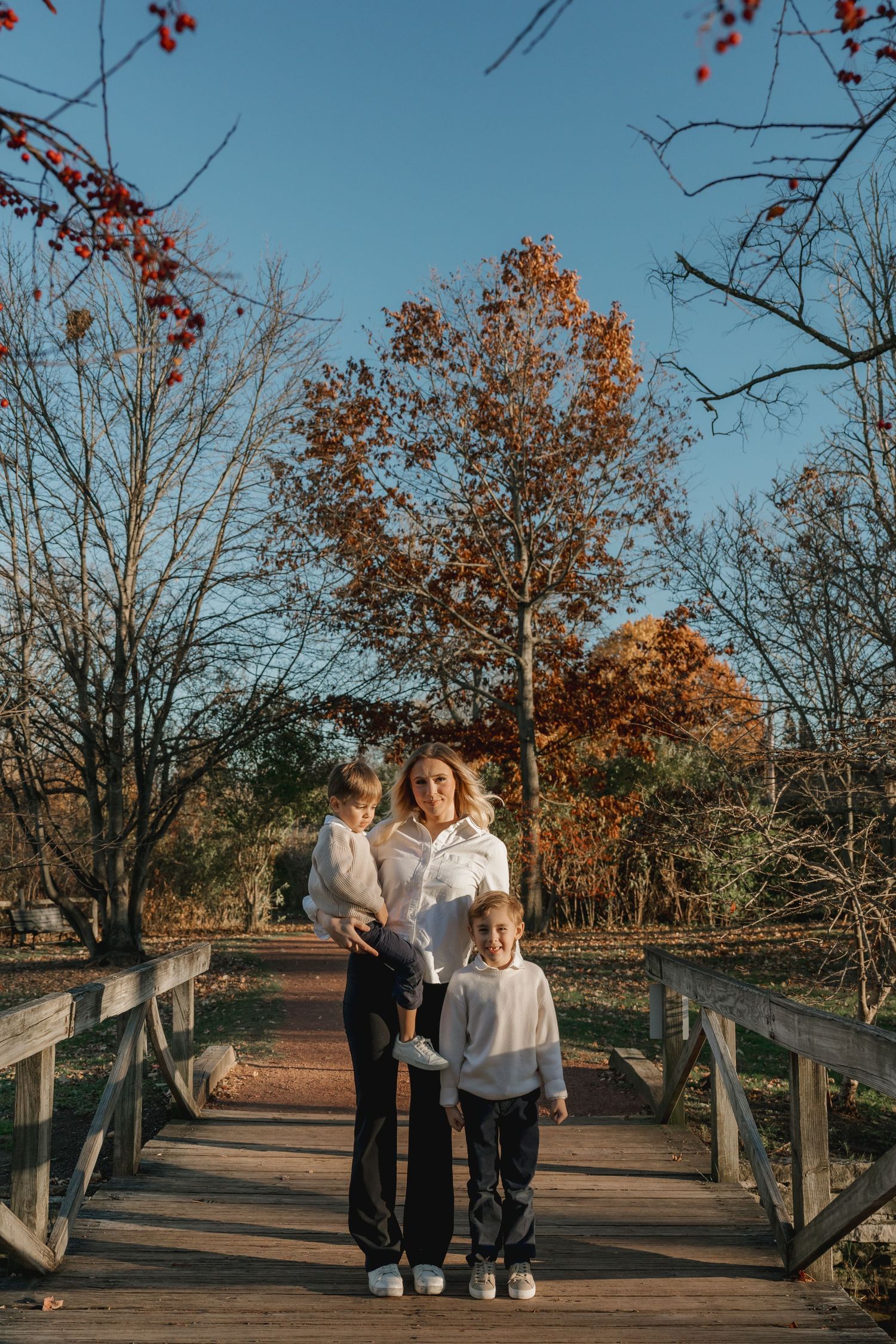 A mother poses with her two children on a wooden bridge surrounded by autumn trees and blue sky.