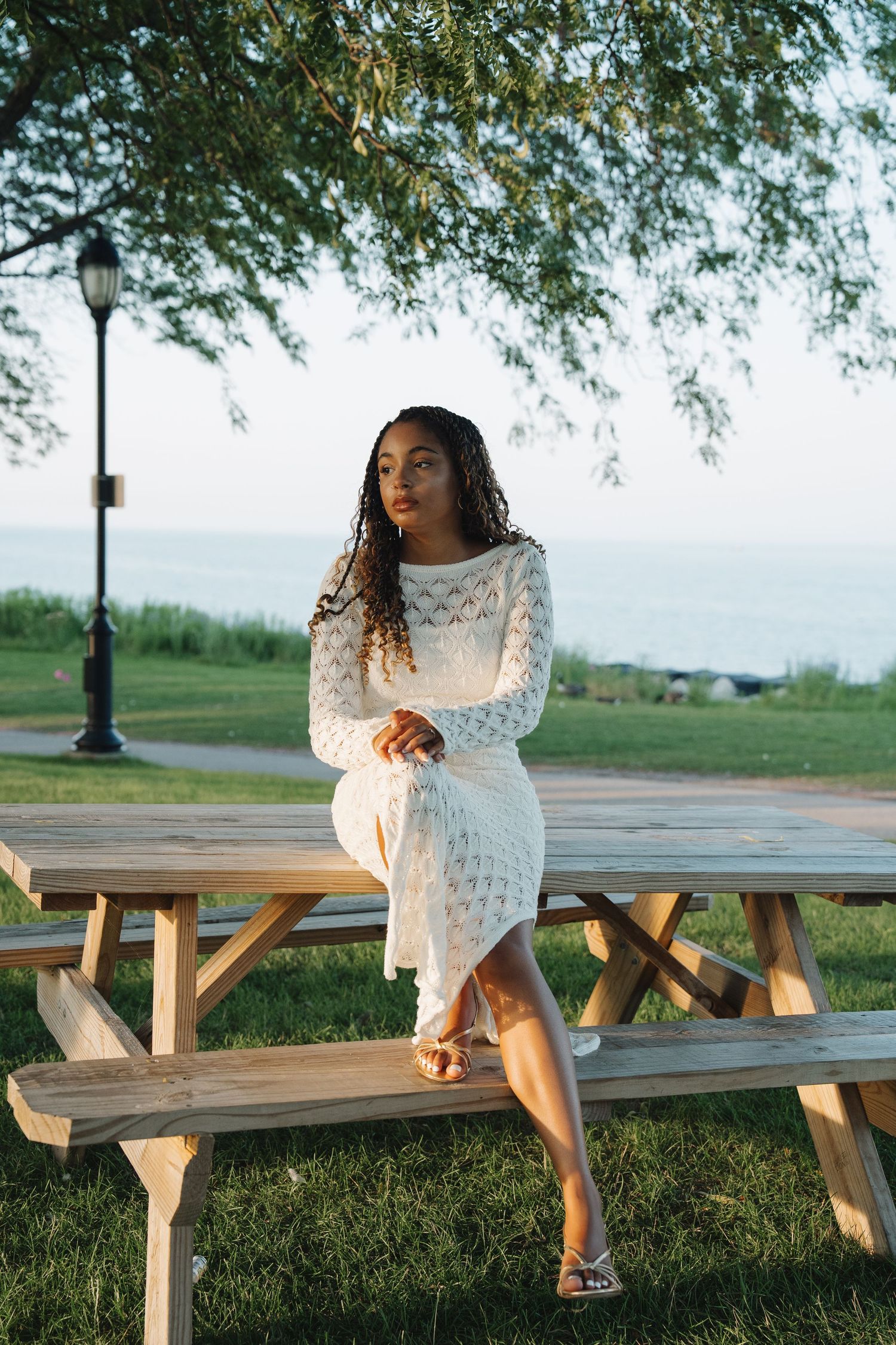 Young woman in white lace dress sitting on wooden picnic table in park near ocean under green tree.