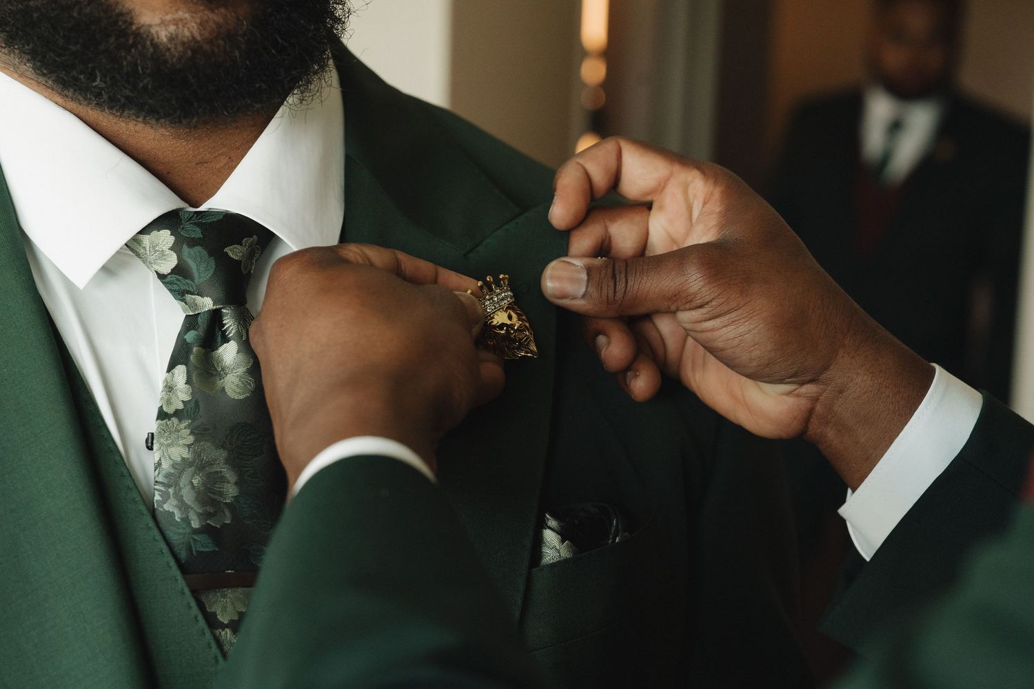 Man in green suit with floral tie being helped with boutonniere by another person wearing a gold ring.