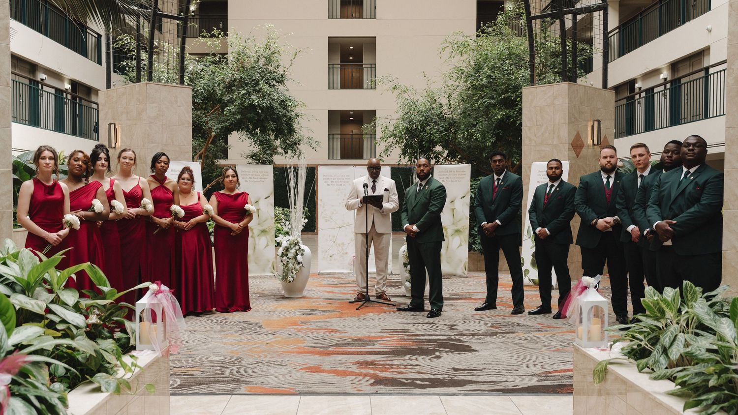Elegant indoor wedding ceremony with bridesmaids in red gowns and groomsmen in dark suits at a hotel atrium venue.