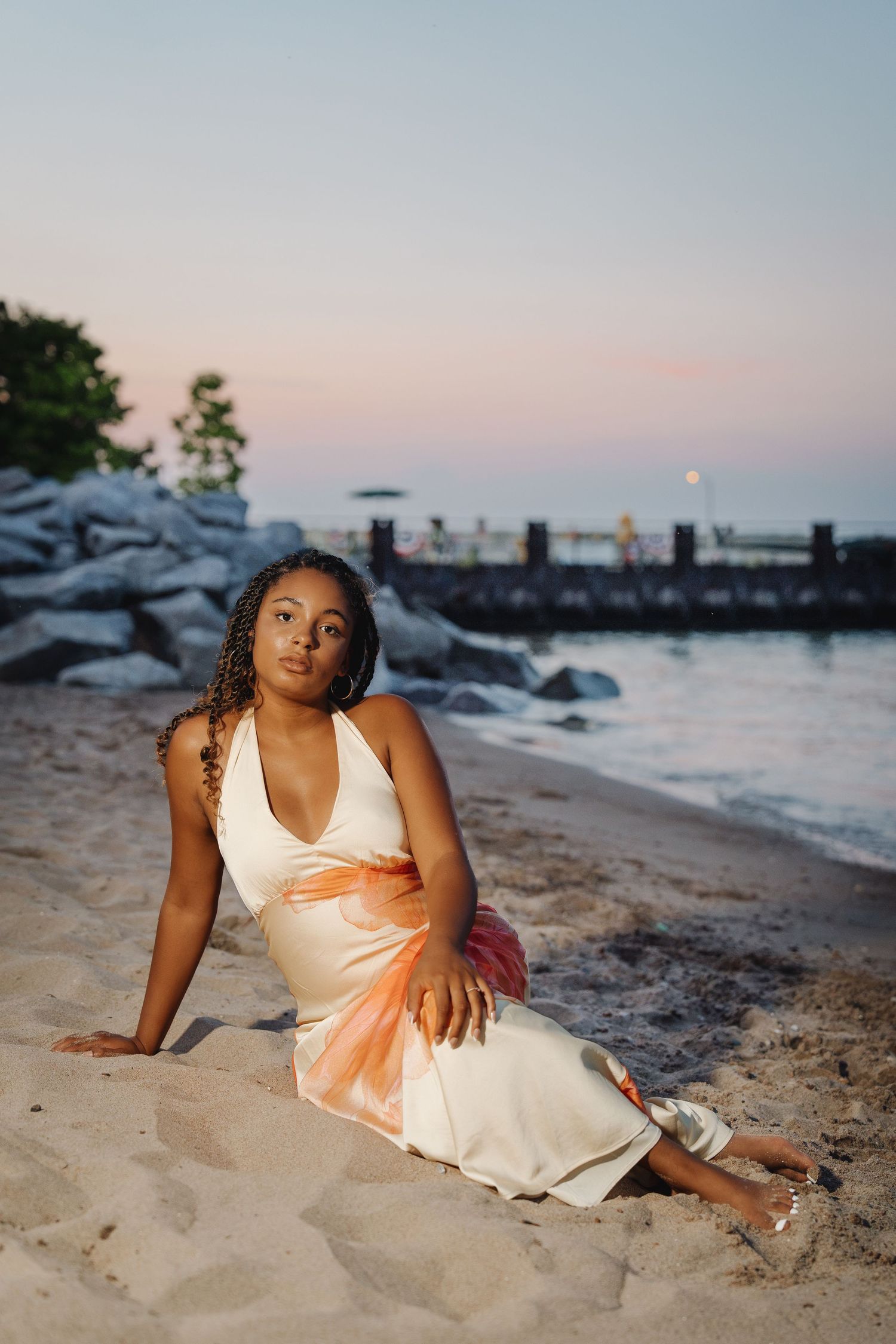 Young woman in white and orange dress sitting gracefully on sandy beach at dusk with rocky shoreline behind her.