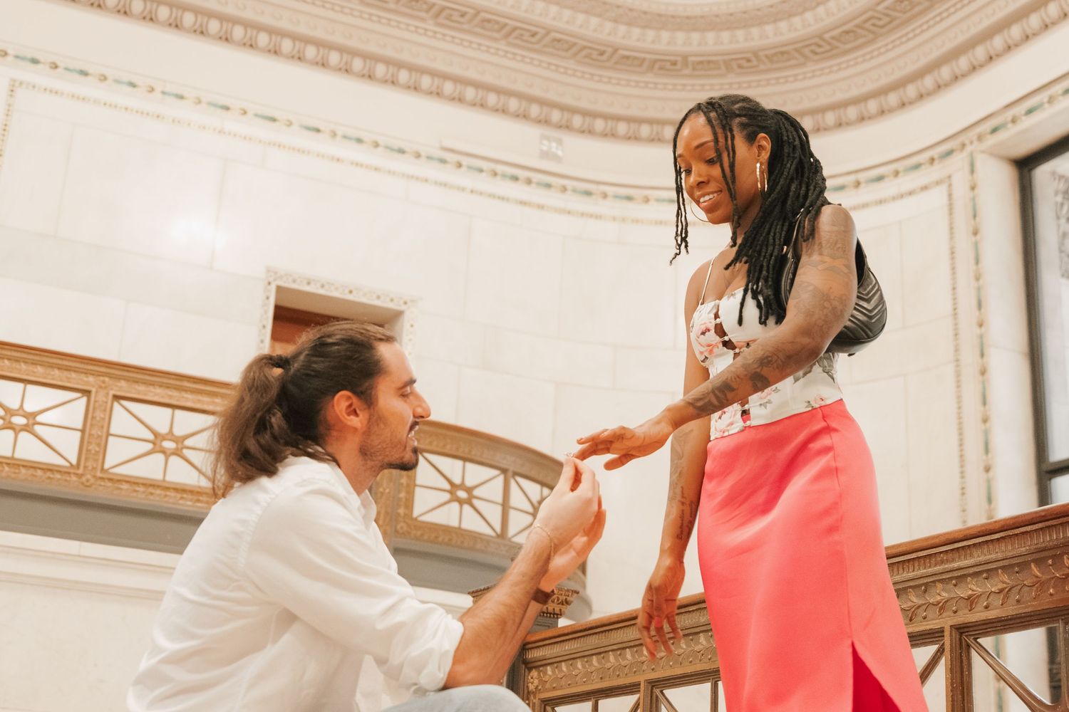 Man kneeling on one knee proposing to woman in pink skirt inside elegant white marble building.