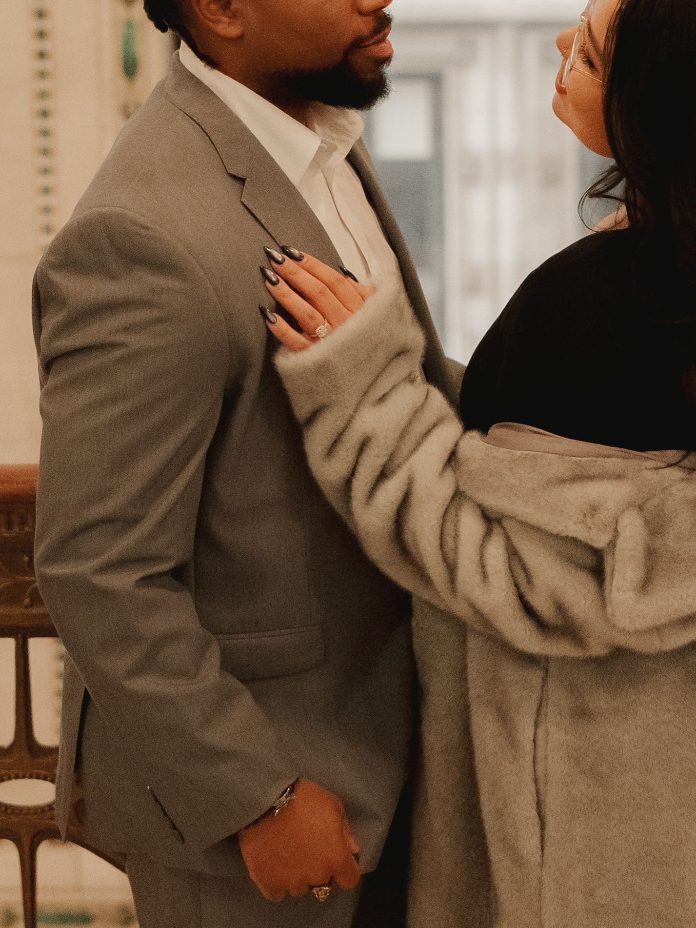 A couple shares an intimate moment, the woman in a gray fur coat resting her hand on the man's chest in a gray suit.