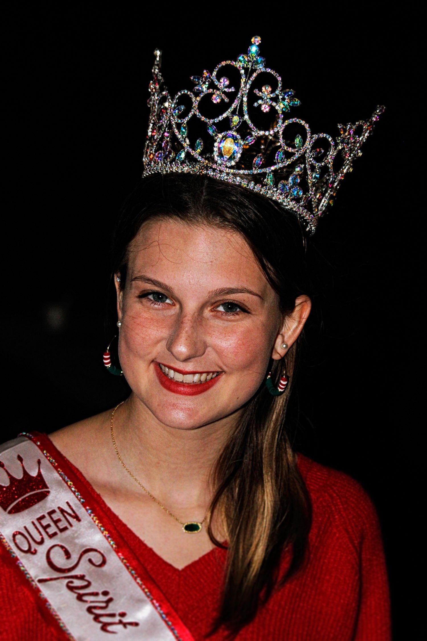 A pageant winner wearing a sparkling crown and red sweater smiles for a portrait.