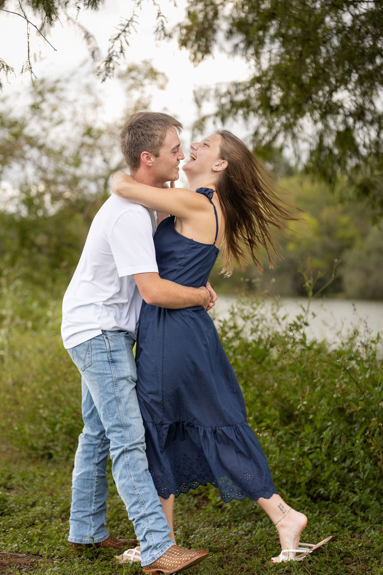 A couple shares a playful moment together near a lake surrounded by greenery.