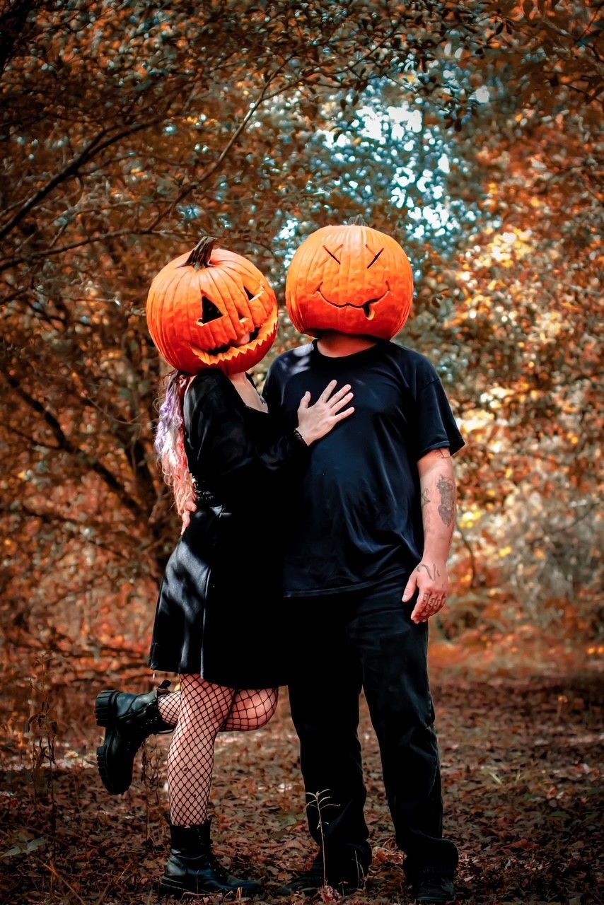 Two people wearing Halloween costumes and pumpkin heads pose together in an autumn forest setting.